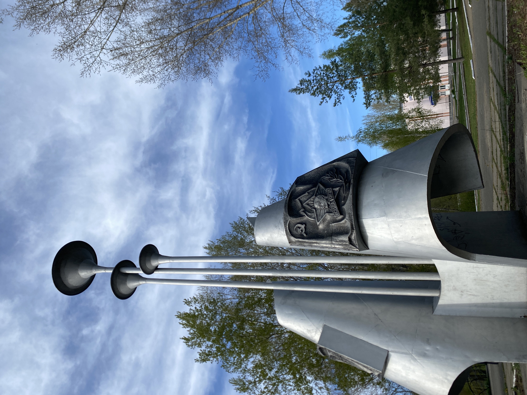 The image shows a low-angle view of the "Three Trumpets" monument in Chaikovsky, Russia, set against a partly cloudy sky. The monument is constructed of metallic materials and features three trumpet-like structures extending upwards. A dark, decorative plaque is affixed to the monument. Trees are visible in the background, and a building and sidewalk are situated to the right of the monument. The photograph appears to be taken from ground level, looking upwards towards the monument and sky.