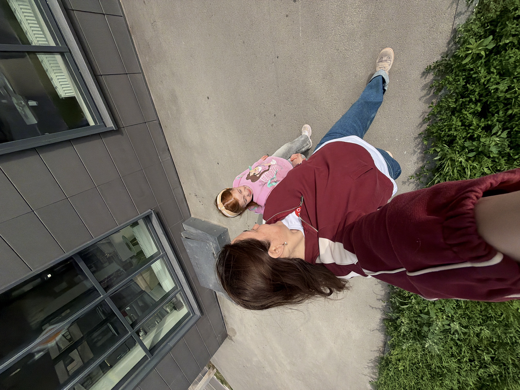 A woman and a young girl are standing on a concrete sidewalk next to a building. The building has dark gray, rectangular facade tiles and large windows. The woman is in the foreground, taking a selfie. She has dark brown hair and is wearing a maroon jacket with white trim over a white t-shirt and blue jeans. The girl is to her right, facing away from the camera. She has red hair with a headband and is wearing a pink long-sleeved shirt with a graphic print and light blue jeans. Both are wearing sneakers. Lush green plants are growing along the edge of the sidewalk. The scene appears to be outdoors, possibly in an urban environment.
