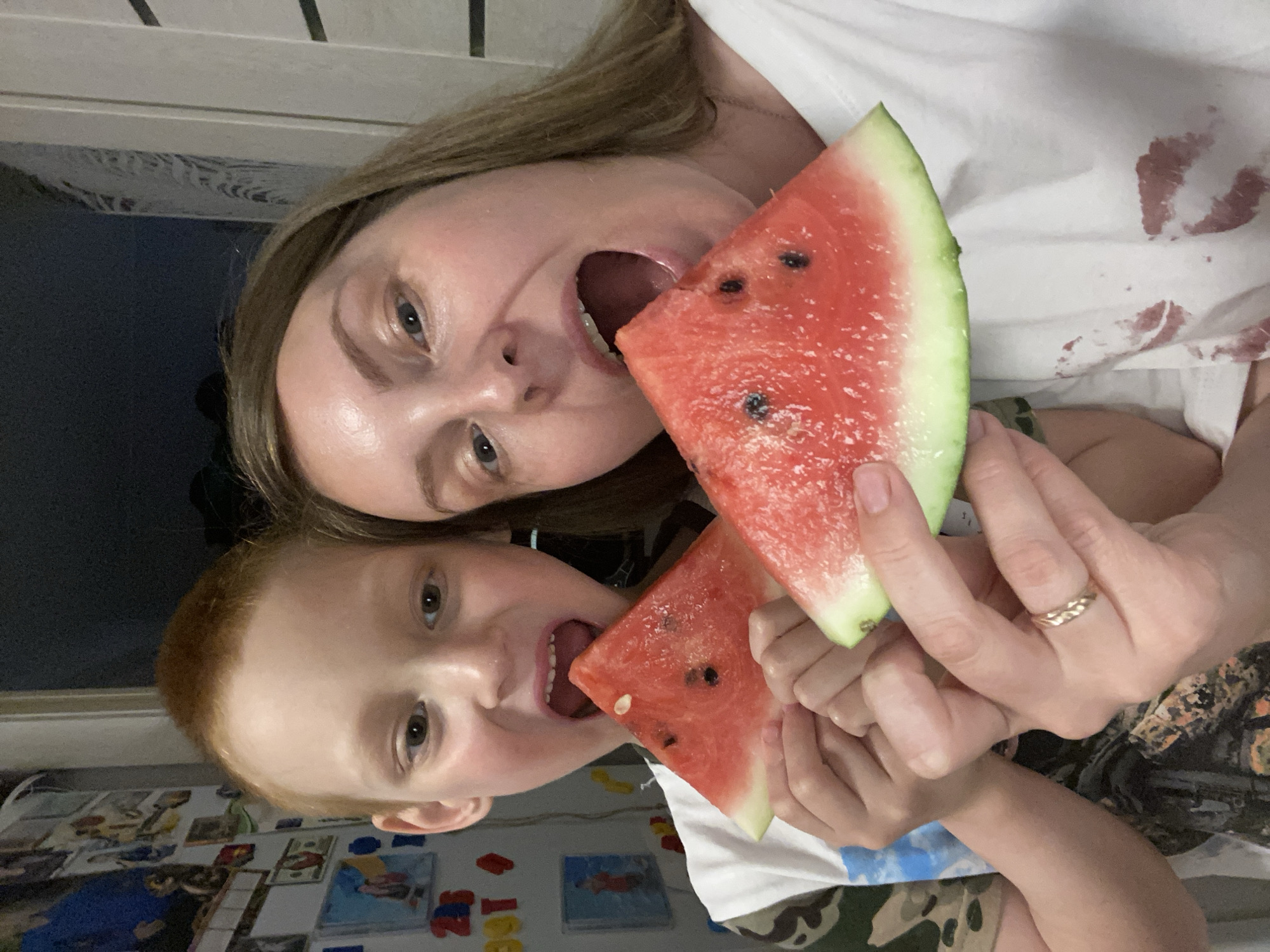 A young woman and a young boy are eating watermelon indoors. They each hold a slice of watermelon in their hands and are biting into it. The woman has blonde hair and is wearing a white shirt with some red stains on it. She has a gold ring on her ring finger. The boy has red hair and is wearing a white shirt with a camouflage pattern on the bottom. The background is a wall with various children's items. It is not possible to confirm whether this is taking place in the city of Noviy, Russia.