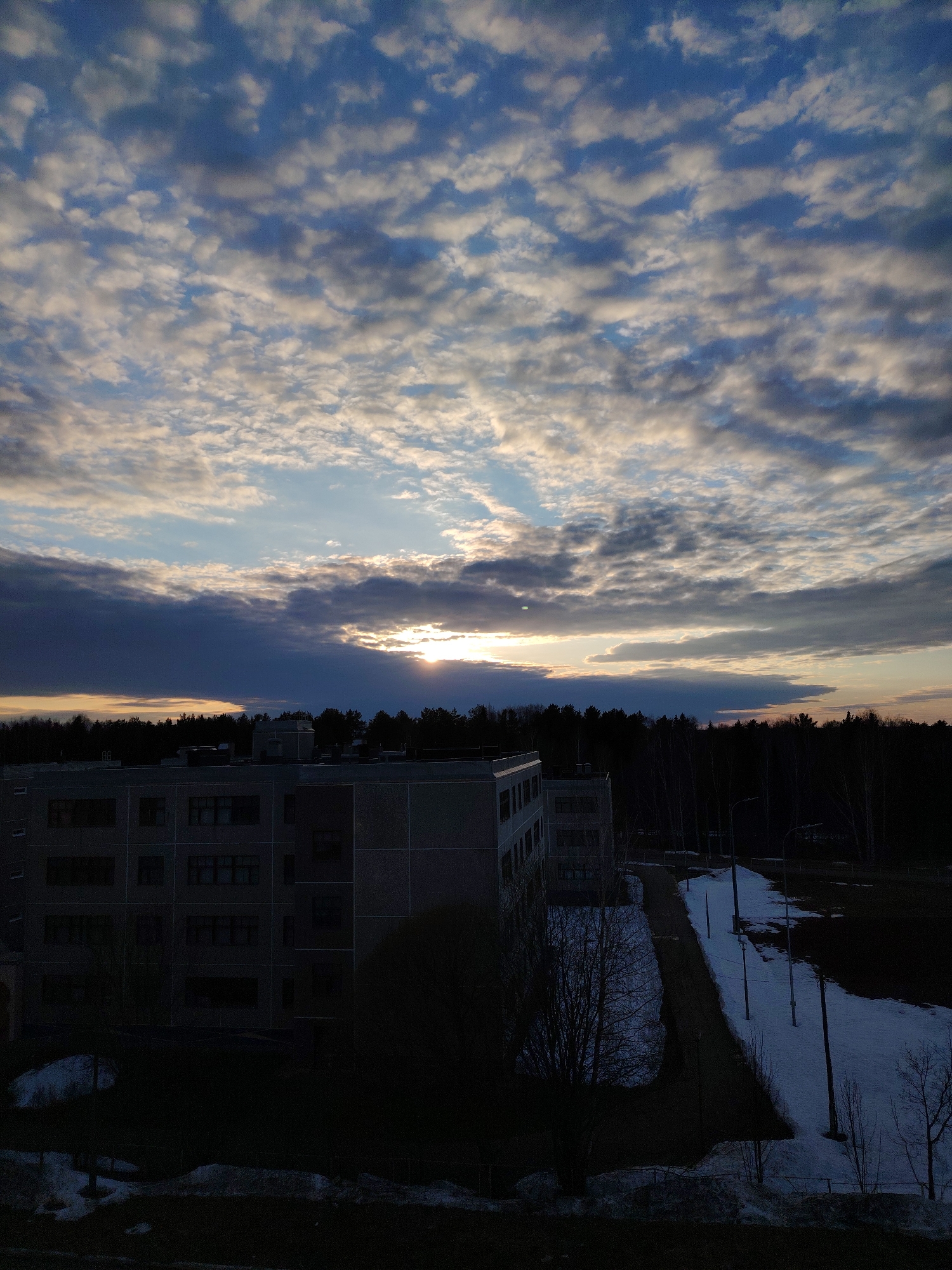 The photograph, taken in what appears to be the city of Марковский, Russia, depicts an urban landscape under a sky filled with altocumulus clouds during a sunset. A large, multi-story building with a flat roof dominates the midground, partially obscured in shadow. The structure appears to be a public or residential building, given the regularity of the window placement. A dense line of trees forms a dark backdrop behind the building, delineating the horizon. A paved walkway runs alongside the building, flanked by patches of snow and sparse vegetation. Several light poles line the walkway. The foreground is mostly dark, with glimpses of grass and melting snow. The sky exhibits a gradient of color, with brighter hues of orange and yellow near the horizon where the sun is setting, filtering through a layer of thicker clouds. The overall atmosphere suggests a transition from winter to spring.