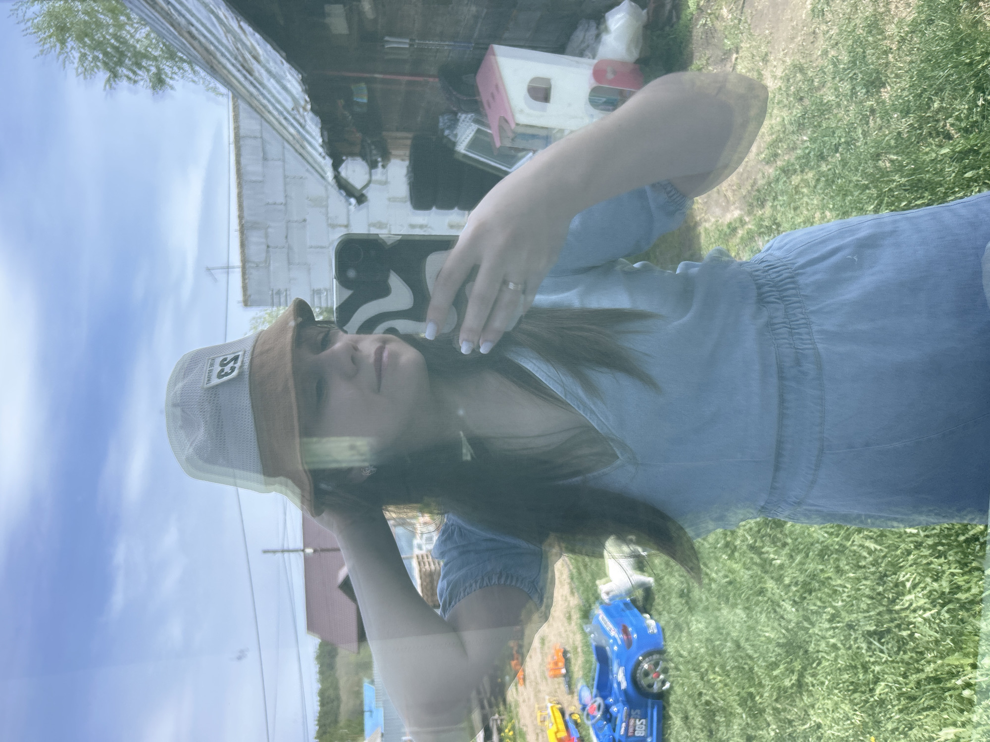 A woman is reflected in a window, holding a smartphone in her left hand. She wears a denim-like dress, a brown and mesh bucket hat with the number "53" on it, and a ring on her left ring finger. Her hair is dark and long. The reflection shows the exterior to include a building with light-colored brick walls and a corrugated metal roof, sky, green grass, and children's toy cars. The location is likely in a residential area in Chaikovsky, Russia.