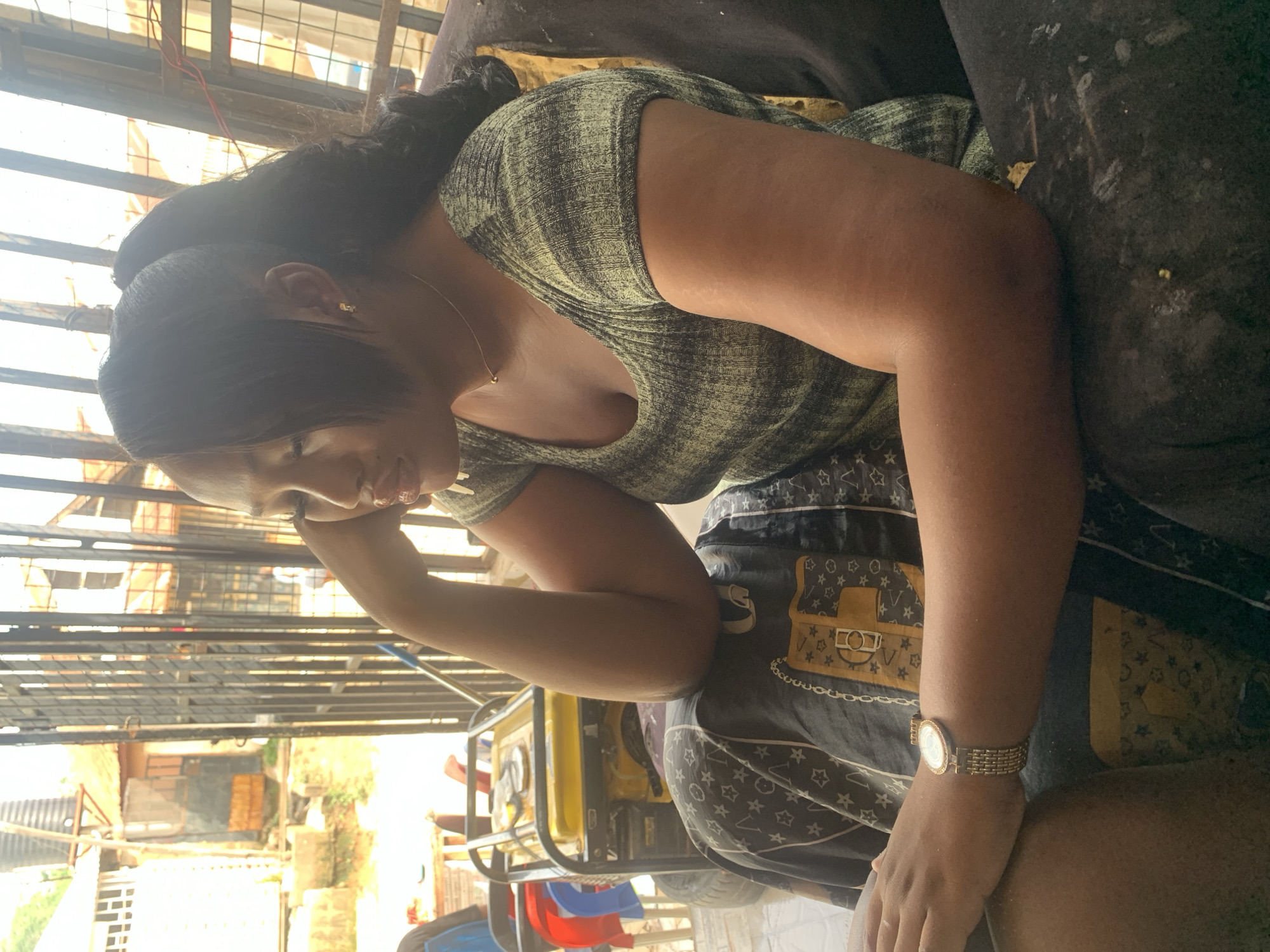 A woman sits indoors on an upholstered surface, wearing a heathered green top, dark patterned shorts, and a gold-toned wristwatch. She rests her face in her hand. Visible through an adjacent window are sections of buildings, foliage, and fencing, suggesting an urban setting. A generator sits near her chair. The location may be in a residential area.