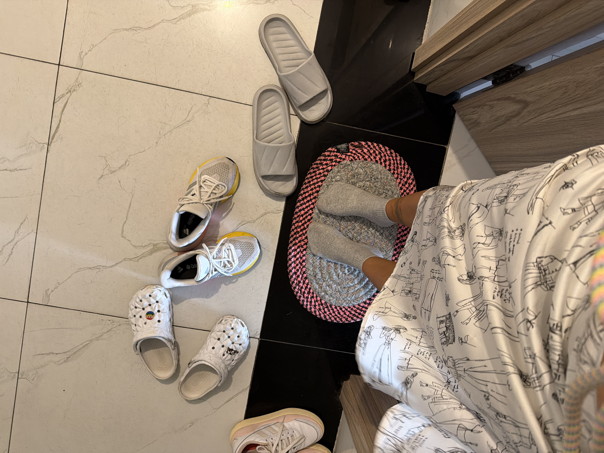 A high-angle medium shot captures a collection of footwear and a person's legs on a tiled floor in what appears to be a domestic setting. The floor is a mix of large, light-colored tiles with faint marble-like veining and a section of glossy black tile. Scattered across the tiles are several pairs of shoes and slippers, suggesting a recent change of footwear or preparation for going out.
On the white tiles, a pair of white Crocs with a perforated design and a small circular colorful emblem on one shoe are visible. Next to them, another white Croc lies on its side. Two pairs of athletic sneakers are also present: one is white with yellow accents on the sole and grey laces, and the other is also white with yellow and orange accents on the sole and white laces.
On the black tiled area, a pair of light grey slide slippers are placed one after another. A circular mat with a pink and grey woven pattern sits on the black tiles, and a person's legs, clad in grey socks, are positioned on it. The person is wearing a loose-fitting garment adorned with a black and white sketch pattern of various figures. The leg closest to the viewer shows a tattoo on the shin.
The scene is indoors, possibly near a doorway indicated by the wooden cabinet on the right. The lighting suggests daytime, but there are no clear indicators of the time of day or weather conditions. The overall impression is one of casual domestic life, with a focus on footwear and a person getting ready or relaxing. No visible text is discernible on the shoes or the person's garment.