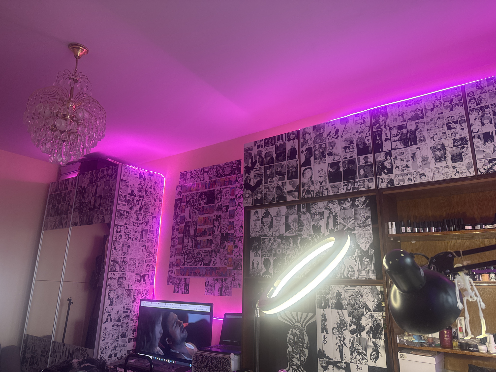The image presents a room interior under a predominantly pink light emanating from LED strips near the ceiling. A chandelier hangs from the ceiling. The walls are covered with manga panels. There's a computer monitor displaying a video, a laptop, a ring light, and a black desk lamp. A wooden bookshelf filled with bottles and other small items is also visible. There is a wardrobe with mirrored panels on the left. The scene appears to be indoors, possibly in a bedroom or workspace. The location is undetermined, and the presence of Moscow, Russia cannot be verified.