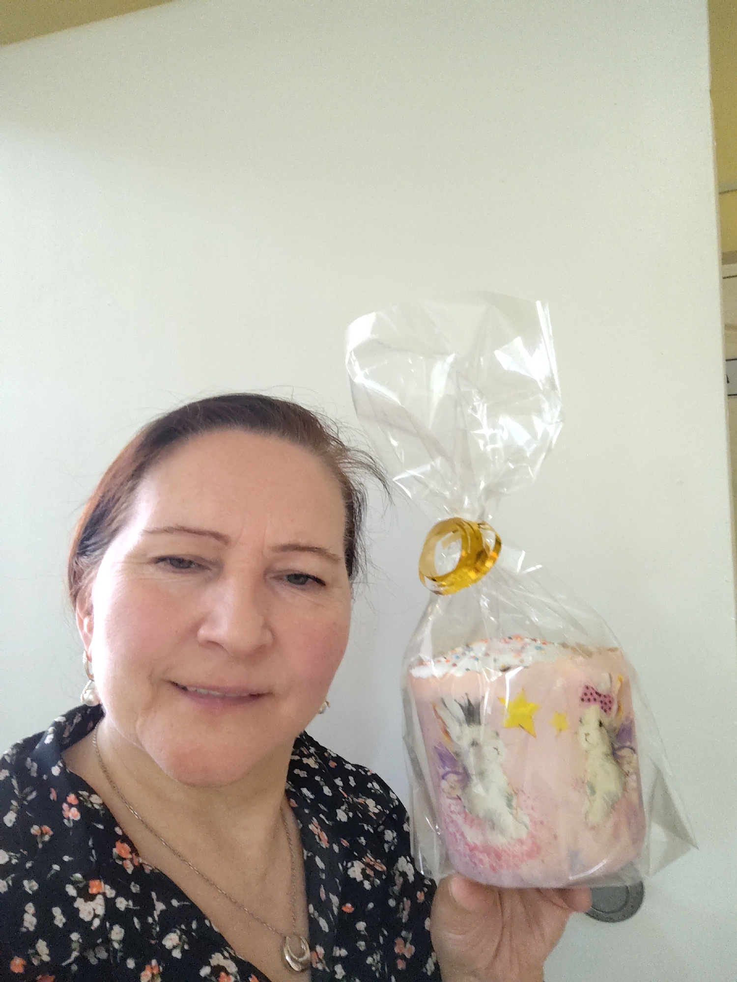 A woman is holding a wrapped Easter cake. The cake is pink with white bunnies on it, covered in sprinkles, and wrapped in clear plastic with a gold tie. The woman has auburn hair and is wearing a floral blouse and jewelry. The background appears to be a white wall and doorframe. It is not possible to confirm the location in Sarapul, Russia from this image.