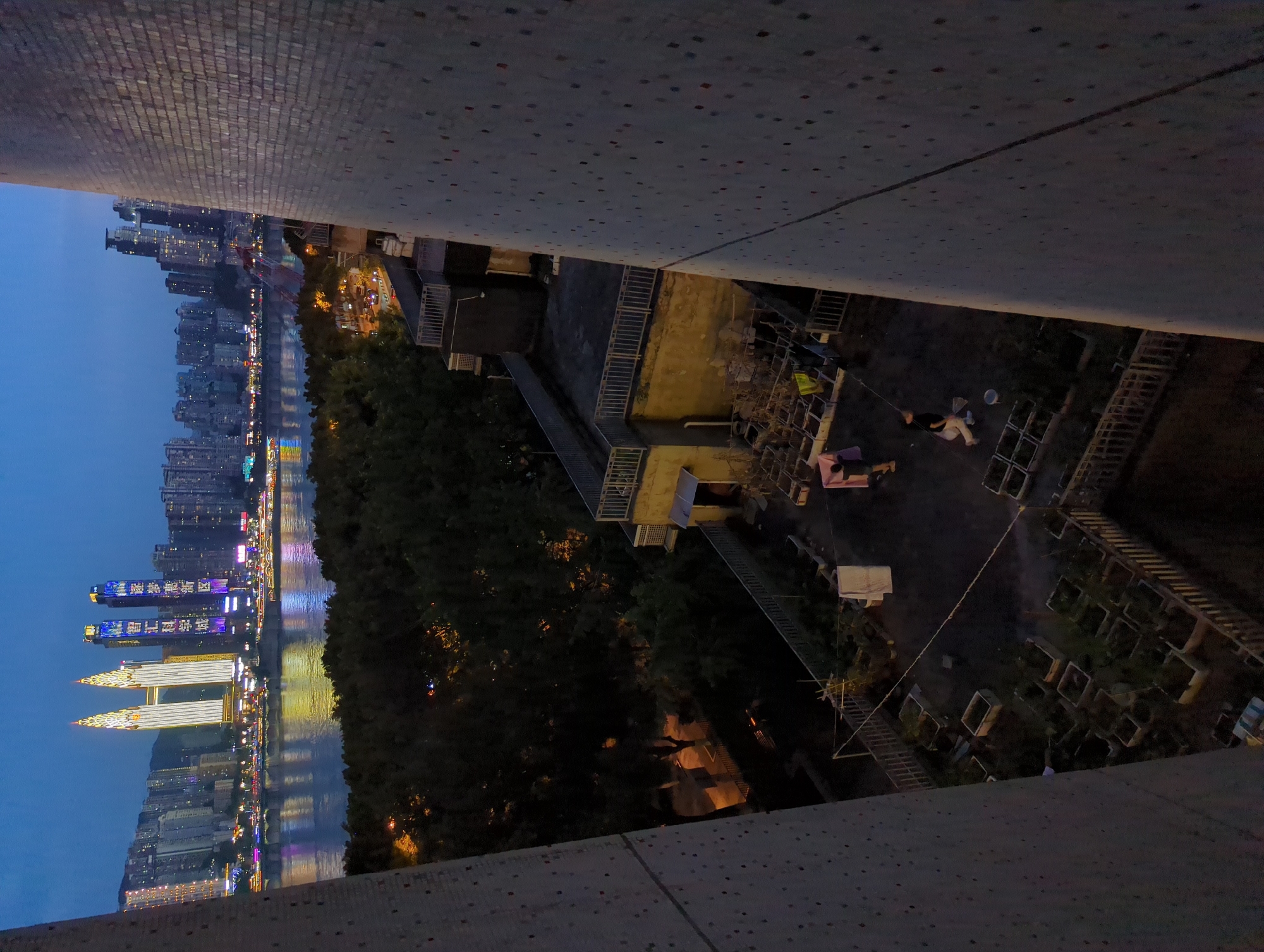 This is a nighttime, high-angle view of a rooftop garden in Yuzhong District, Chongqing, China. In the foreground, two large, textured building facades flank the scene. The rooftop is filled with numerous square planters containing small plants. A clothesline stretches across the garden, with several items of laundry hanging on it, including a white sheet. Two individuals are present on the rooftop; one, a person wearing a dark shirt and light pants, is walking across the garden, and another person in the background is partially obscured. Adjacent to the garden are several low-rise buildings with balconies and railings. In the background, across a wide body of water, the illuminated cityscape of Chongqing stretches along the waterfront. Notable are several brightly lit skyscrapers, including two prominent twin towers with ornate lighting, and a Ferris wheel structure. The sky is a deep twilight blue.
