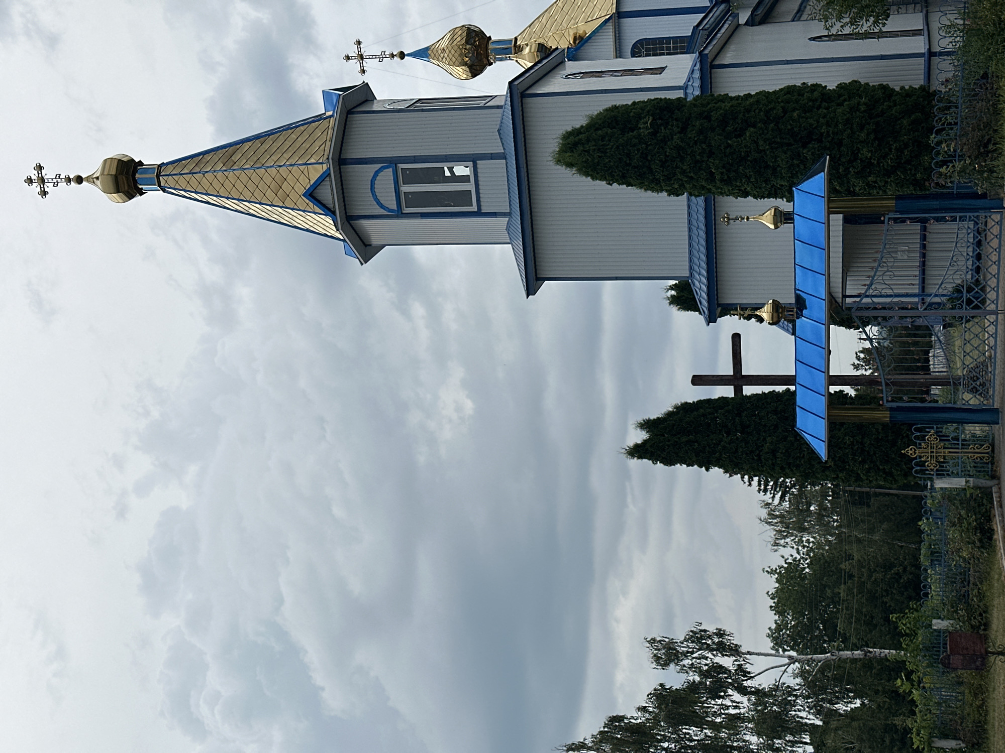 A full, eye-level, outdoor shot shows a predominantly white church with blue trim and gold domes under a cloudy sky. The church is to the right of the frame, with its prominent bell tower topped by a gold dome with a cross, and a smaller gold dome visible to its right. A tall, dark wooden cross stands to the left of the church's entrance, which is framed by a blue awning and a decorative metal gate with gold accents. The church is flanked by tall, dark green evergreen trees, and in the background, a line of deciduous trees with white trunks are visible. The ground is covered in dense green foliage and a low metal fence.