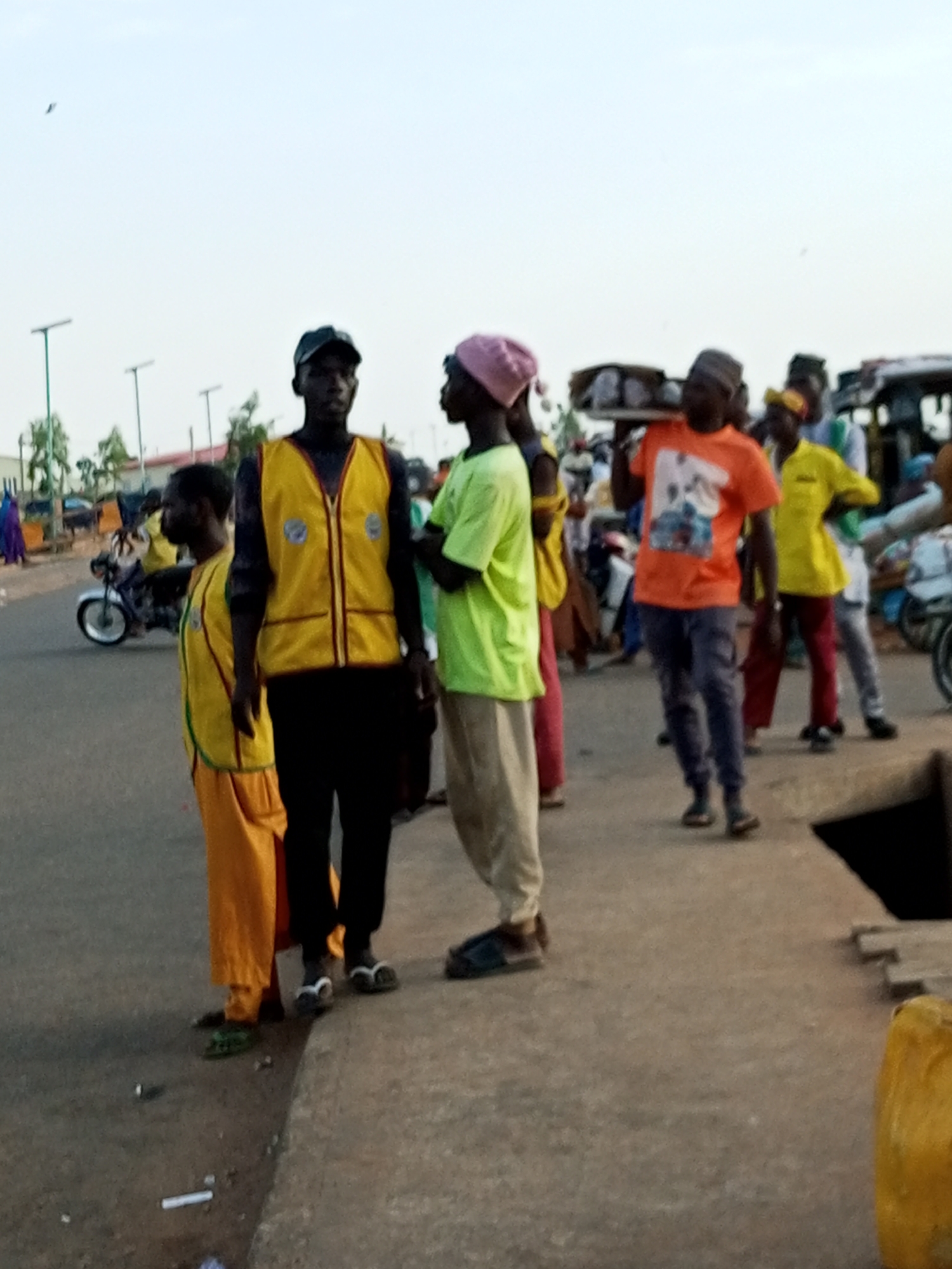 A group of people are standing and walking on a paved road. In the foreground, two men in yellow vests are facing the camera. The man on the left is wearing a black cap and a yellow vest over a black shirt and black pants. He is looking to his left. The man on the right is wearing a black cap and a yellow vest over a black shirt and black pants. He is looking to his right. To their right, a man wearing a pink headscarf and a green shirt is walking away from the camera, and another man is walking away from the camera carrying a box on his shoulder, wearing an orange t-shirt. In the background, there are motorcycles and people walking. The environment appears to be an outdoor market or street scene in Gombe, Nigeria, with a clear sky.