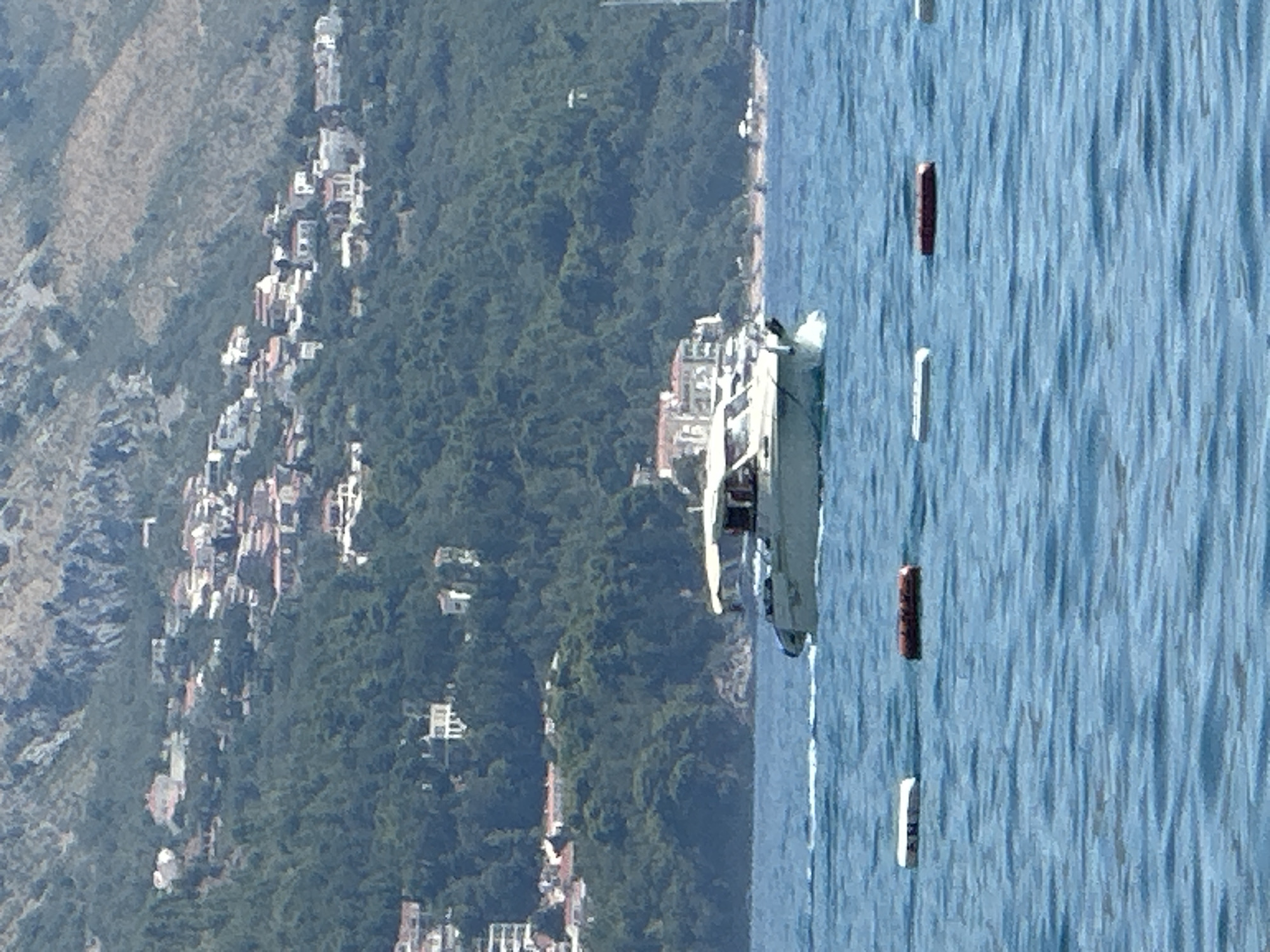 The image captures a scenic view of the coastline in Boreti, Montenegro. In the foreground, calm blue water stretches across the frame, with several buoys floating on the surface, marking a swimming area. A white and grey motorboat is in motion, leaving a slight wake behind it, moving from left to right across the water. In the middle ground, a densely forested hillside rises steeply, dotted with numerous houses and buildings with red-tiled roofs, characteristic of Mediterranean architecture. These structures appear to be built into the slope, creating a terraced effect. The background features a rugged, rocky mountain range, partially covered in sparse vegetation, suggesting a mountainous terrain overlooking the sea. The sky is a pale, washed-out blue, indicating a bright, sunny day. The overall impression is one of a coastal town nestled against a dramatic natural landscape.