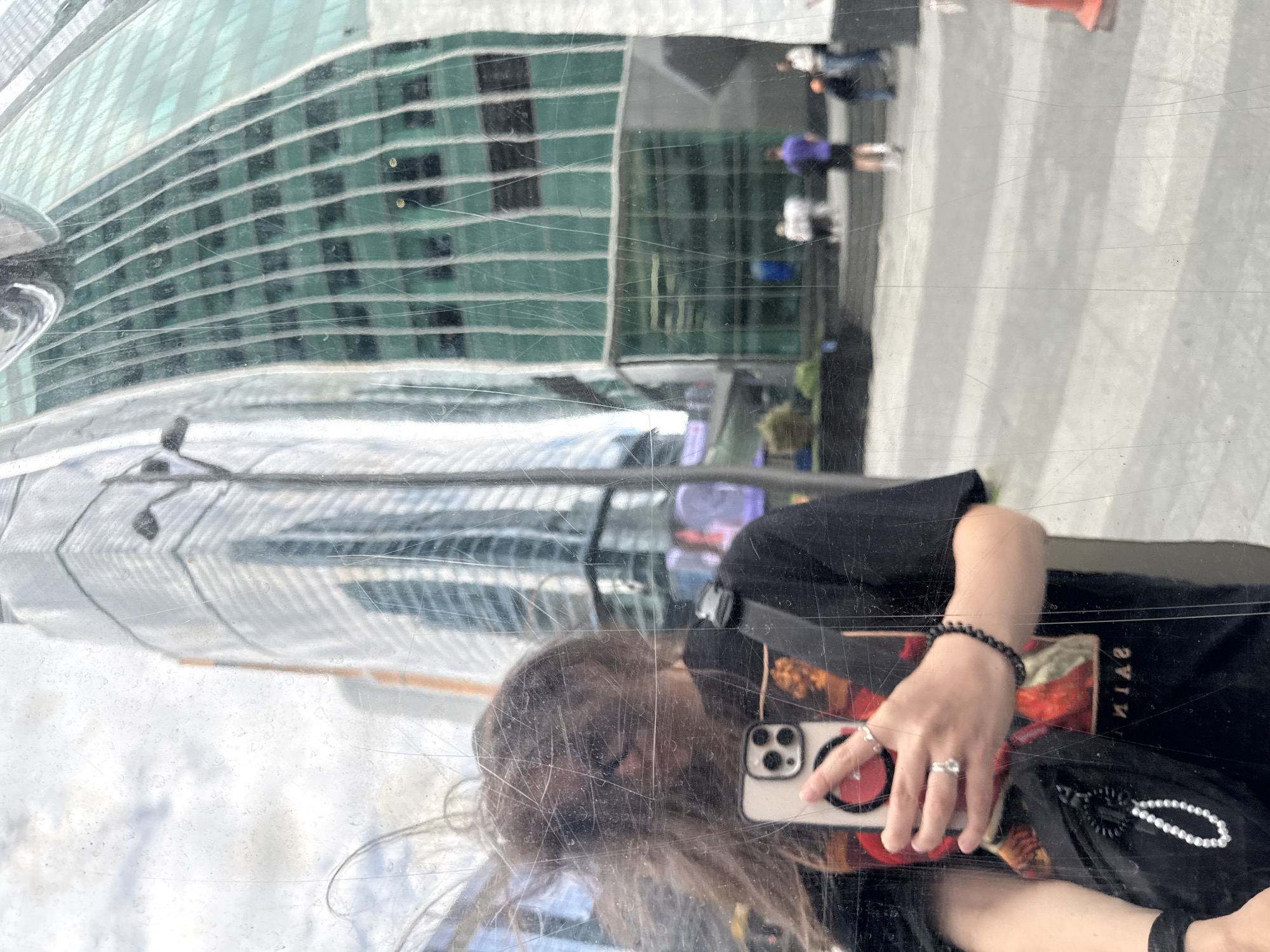 The image is a reflection of a person taking a selfie in a glass surface. The reflection shows the person holding a smartphone, wearing a black t-shirt, a black bracelet and a ring. The reflection also shows a large building with a green glass facade and other people walking on a sidewalk. The city is Moscow.