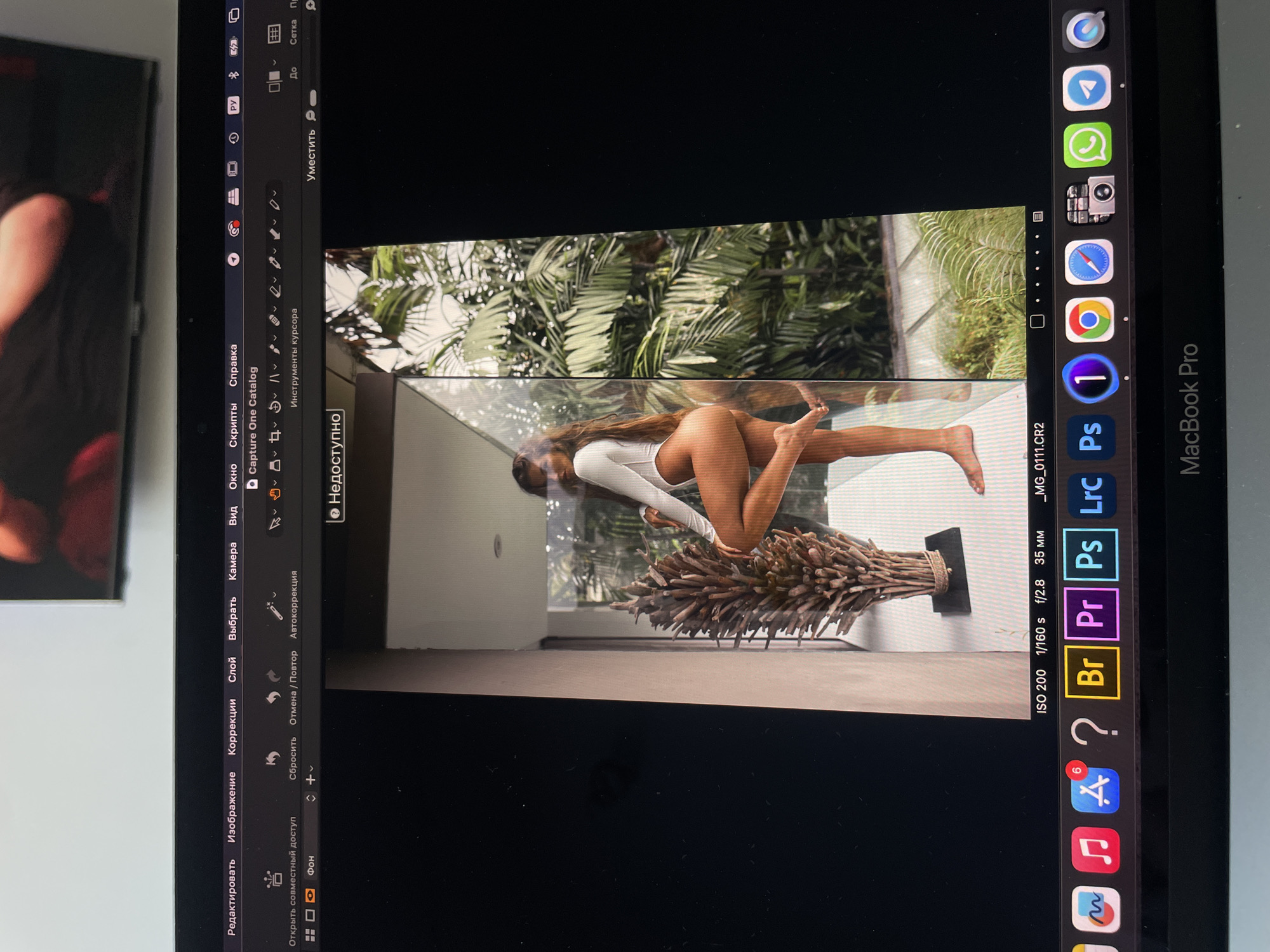 A photograph taken of a Macbook Pro screen shows a photo editing software interface. The main window displays a vertically oriented image of a woman in a white swimsuit striking a pose on what appears to be a glass surface. Below her is a decorative arrangement of bundled wooden sticks. The background includes blurred greenery, possibly a lush garden or forest. On the left side of the screen is the photo editing software's menu and toolbars, with labels in Russian. The right side of the screen shows the MacOS dock containing icons for various applications. The EXIF data visible in the bottom right indicates the image was captured at ISO 200, 1/160 s shutter speed, f/2.8 aperture, and a 35mm focal length. There is no indication of the location being Kazan, Russia.