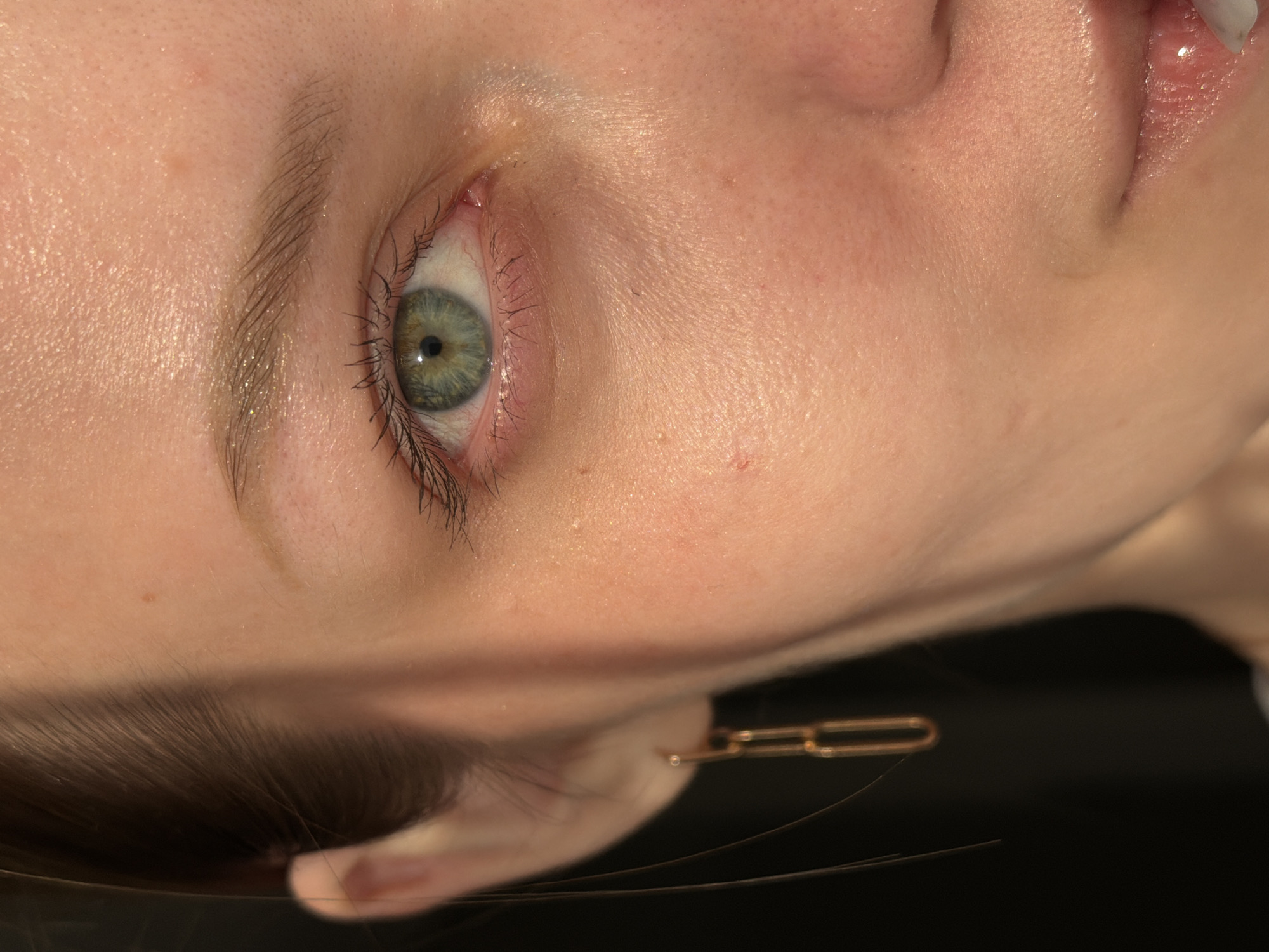 A close-up of a person's face angled so that the left eye is the most prominent feature. The eye has a mixture of green and gray hues. The skin has a dewy appearance. A gold chain earring is visible near the ear. Hair is visible near the ear and forehead. The background is a dark, indistinct color. The location and city of Reshetnikovo, Russia are not discernible from the image.