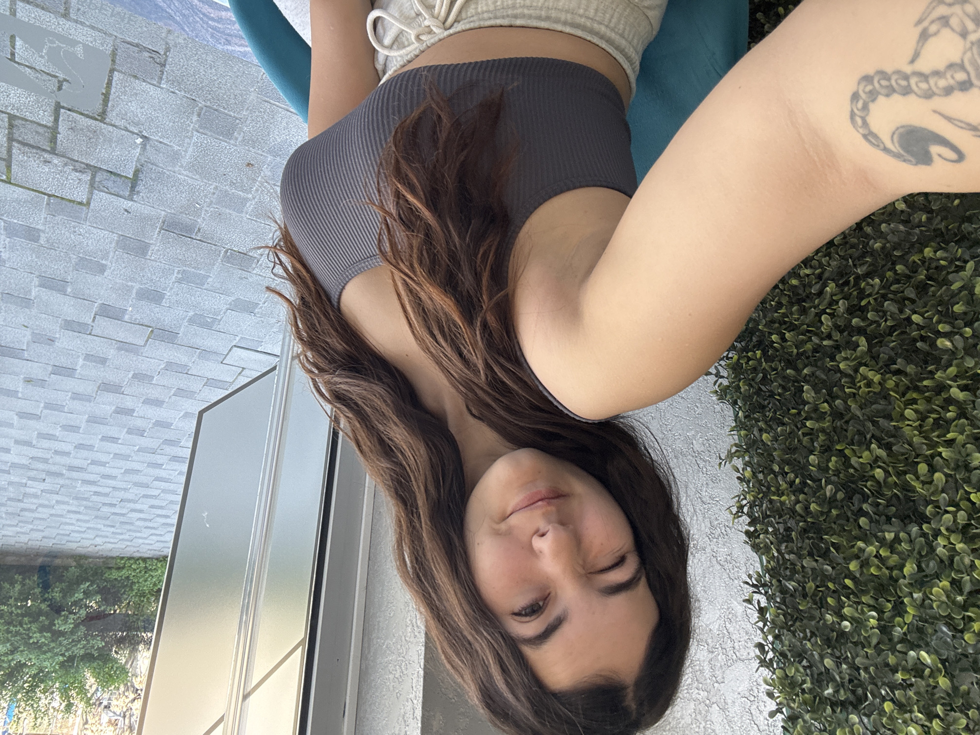 A woman is pictured in a selfie. She has long brown hair, is wearing a gray tank top and light-colored bottoms, and is winking at the camera. She has a tattoo on her upper arm. She is sitting on a blue cushion. To her left is an aluminum frame and glass, reflecting trees and sunlight. To her right is a wall covered in green foliage. The ground is paved with gray interlocking stones. There is no evidence in the image to confirm the location is in Plovdiv, Bulgaria.