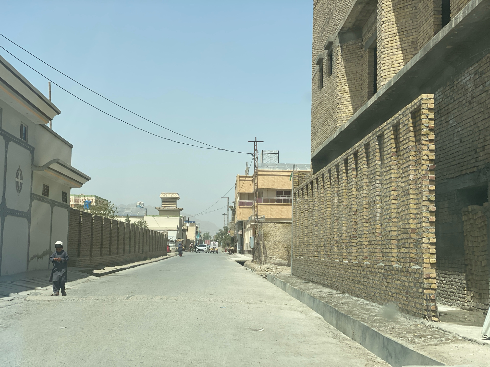 The image depicts a street scene in the city of Kandahar, Afghanistan, under a clear, hazy sky. A young boy wearing a white cap and a dark shalwar kameez walks on the left side of the paved road. Adjacent to him is a pale-colored building with decorative grey detailing and a flat roof. The street is lined on both sides with brick walls. On the right side of the street, a large, unfinished brick building with multiple levels and small, recessed windows dominates the foreground. Its facade is constructed of light brown bricks, and a concrete ledge runs horizontally across its second story. A concrete drainage ditch runs along the curb. In the distance, other buildings, some under construction, are visible, including structures with solar panels on their roofs. A few vehicles, including a white car and a tuk-tuk, are present further down the street. A woman carrying a load on her head is seen walking in the distance. Overhead, power lines stretch across the sky, supported by wooden poles. The overall impression is of a developing urban area with ongoing construction and daily life.
