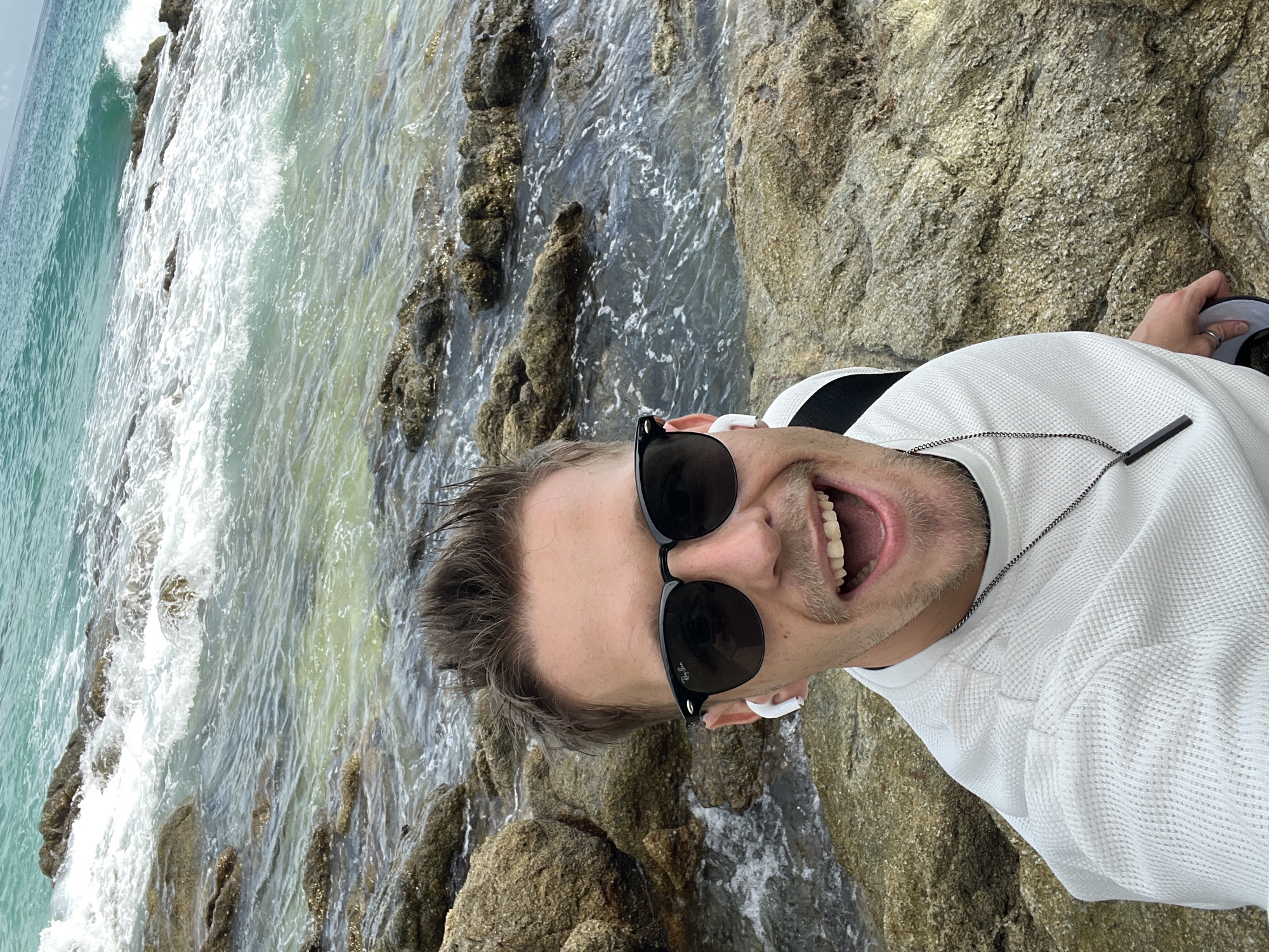 A close-up, selfie-style photograph taken by a man at a rocky coastline in Choeng Thale, Thailand. The man, with short brown hair, is wearing black sunglasses, white earbuds, and a white crewneck shirt with black accents on the shoulders. He has an open-mouthed expression, revealing his teeth. He is adorned with a silver chain necklace with a black pendant. The background shows the ocean with waves crashing against rocks, and the clear turquoise water indicates a shallow area. The overall environment is natural and coastal.