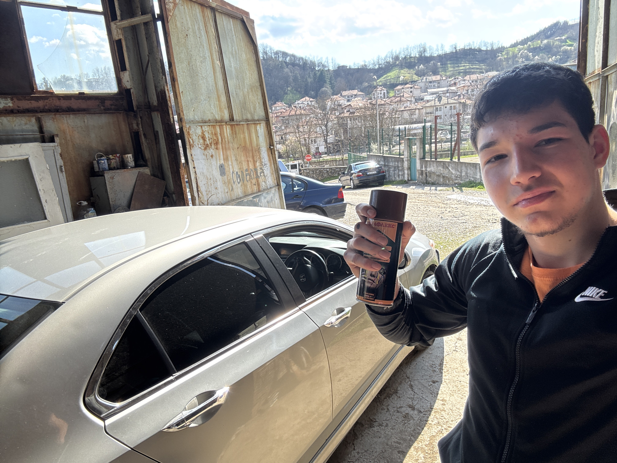 A young man is taking a selfie outdoors, holding a can of car detailing spray in his right hand. He is wearing a black Nike zip-up jacket over an orange shirt. Behind him is a silver sedan, which appears to be the focus of his attention. In the background, a rural scene unfolds with houses dotting the hillside. This area is likely Zlatograd, Bulgaria due to the architectural style and landscape. The photo is taken near a structure made of metal and possibly wood. There are other cars parked further away, but they are not the main subject. The lighting indicates it is daytime, with some areas brightly lit while others are in shadow.