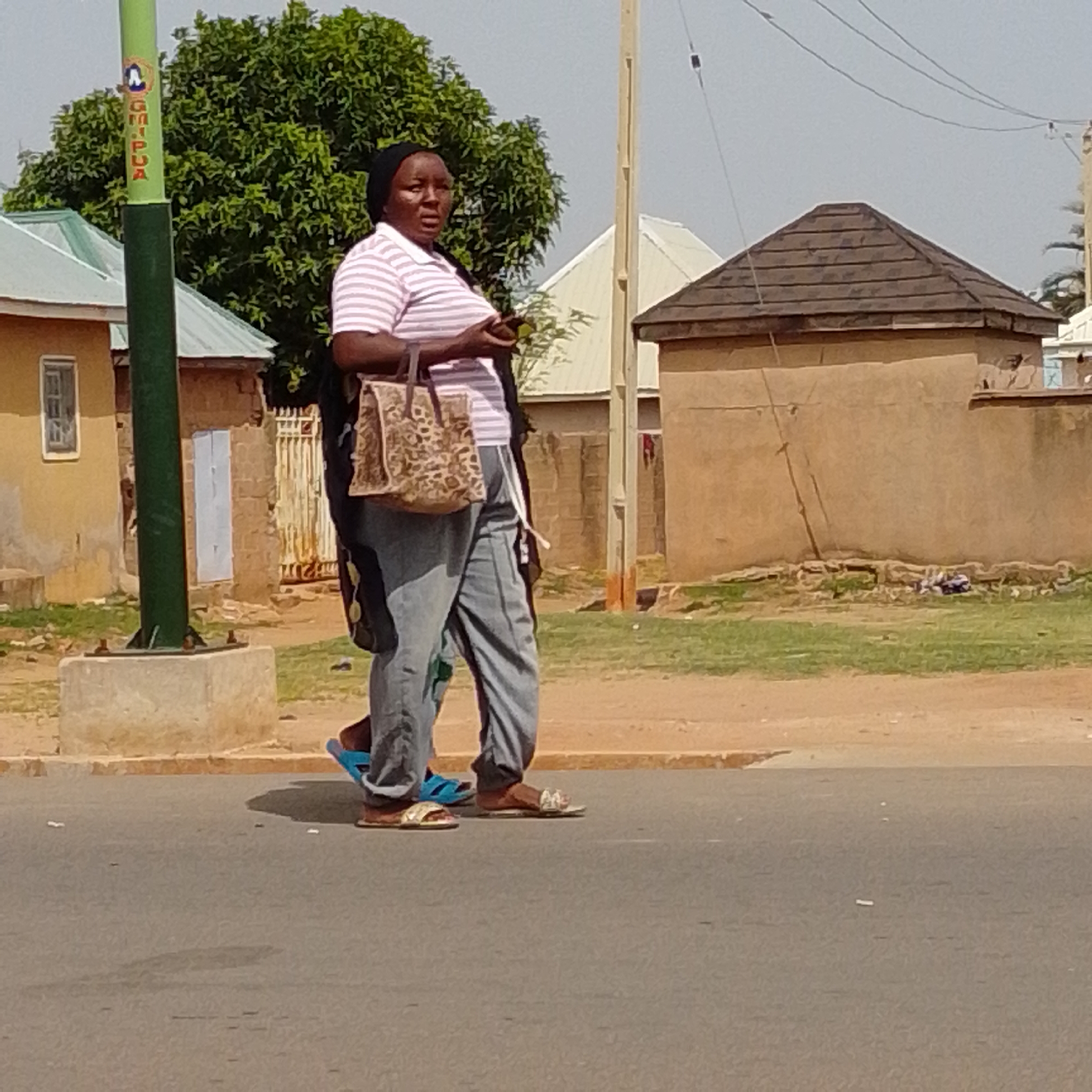 A woman wearing a black headscarf, a pink and white striped t-shirt, and light grey pants is walking on a paved road. She is carrying a light brown patterned handbag on her right arm. She is wearing light brown sandals on her right foot and a blue sandal on her left foot. Her left leg is bent at the knee as she walks. Another person, only their feet and lower legs are visible, is walking to her left. They are wearing blue flip-flops on their left foot and light brown sandals on their right foot. The environment includes a green pole with text on it to the left, a yellow building with a window, and a brown building with a dark brown, hipped roof behind them. The ground is dirt and grass, and there is a concrete curb. The scene is outdoors in what appears to be a residential or commercial area in the city of Gombe, Nigeria, with a clear sky overhead.