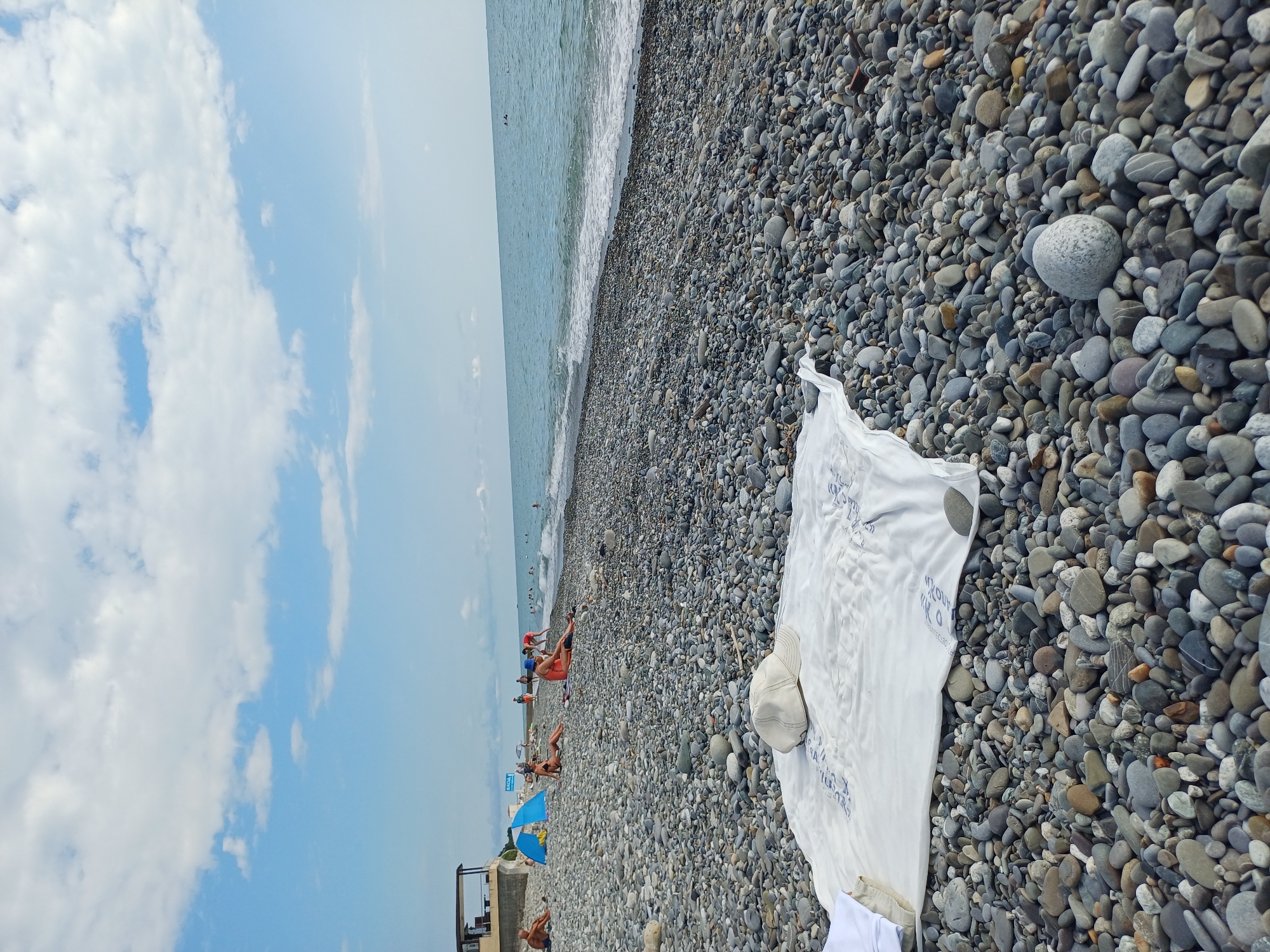 The image depicts a sunny day at a pebble beach, likely in Sochi, Russia. The sky is a bright blue with scattered white clouds. The foreground is dominated by a dense covering of smooth, gray, and brown pebbles, interspersed with larger stones. A white cloth, possibly a beach towel or sheet, is spread out on the pebbles in the middle ground, with some faint blue text printed on it. A light-colored baseball cap rests on the cloth.
In the background, the gentle waves of the sea create a foamy white line as they approach the shore. A few people are visible in the water and on the beach, appearing relaxed and enjoying the warm weather. Some are lying on the pebbles, while others are seated or standing. There are also some blue beach umbrellas scattered along the shoreline, indicating a popular spot for sunbathing and recreation. A small, dark-colored structure, possibly a lifeguard station or changing room, is visible on the far left of the frame. The overall mood is one of leisure and relaxation.