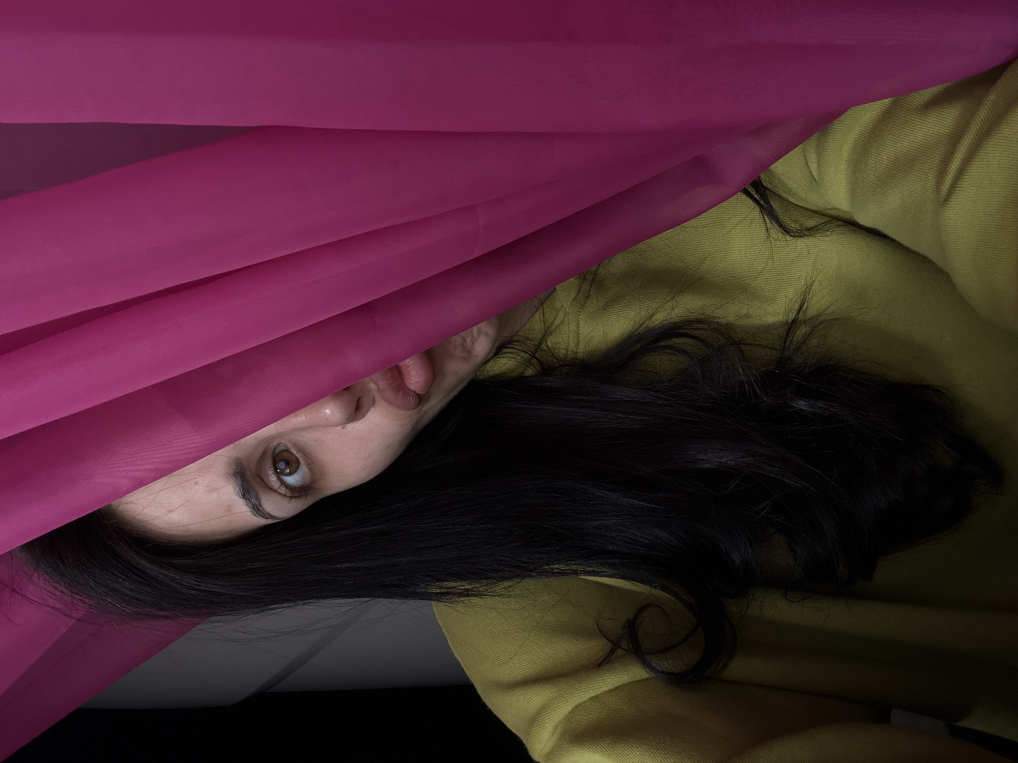 The image shows a close-up of a person playfully peeking from under a vibrant magenta fabric. The person, appearing to be female, is positioned diagonally across the frame, with her head oriented towards the upper left. Her left eye, part of her nose, and mouth are visible peeking from under the sheer fabric. She has a brown eye and long, dark hair that spreads across her shoulder and a chartreuse or lime green garment she is wearing, possibly a sweater or hoodie. Her expression is one of curiosity or playfulness, with her mouth slightly open and a hint of her tongue visible.
The magenta fabric creates soft folds and shadows, dominating the left and upper portions of the image. The green garment covers the right side, while a darker area is visible in the bottom right corner, suggesting an indoor setting. The lighting is soft and diffused. There is no visible text.
Location context: Votkinsk, Russia.