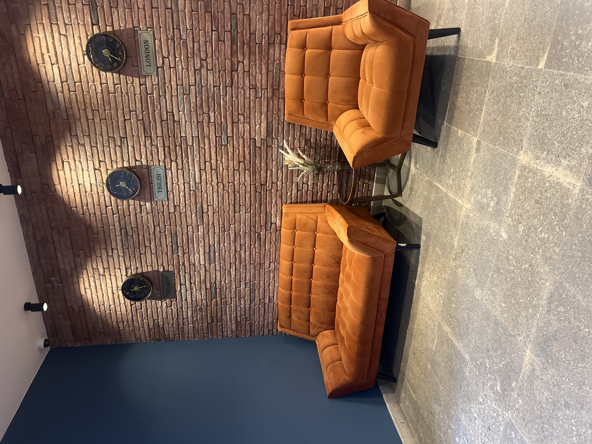 The image shows a waiting area with two orange upholstered chairs and a small round glass-top table with a vase of dried grass. The wall behind the seating is made of exposed brick and is adorned with three clocks, each labeled with a city name: TEL-AVIV, TBILISI, and LONDON. The floor is tiled with large, grey stone-like tiles. The overall scene suggests a modern and stylish waiting room, possibly in a hotel or office. The presence of the clock labeled TBILISI indicates that this location is in Tbilisi, Georgia. The room appears to be empty of people.