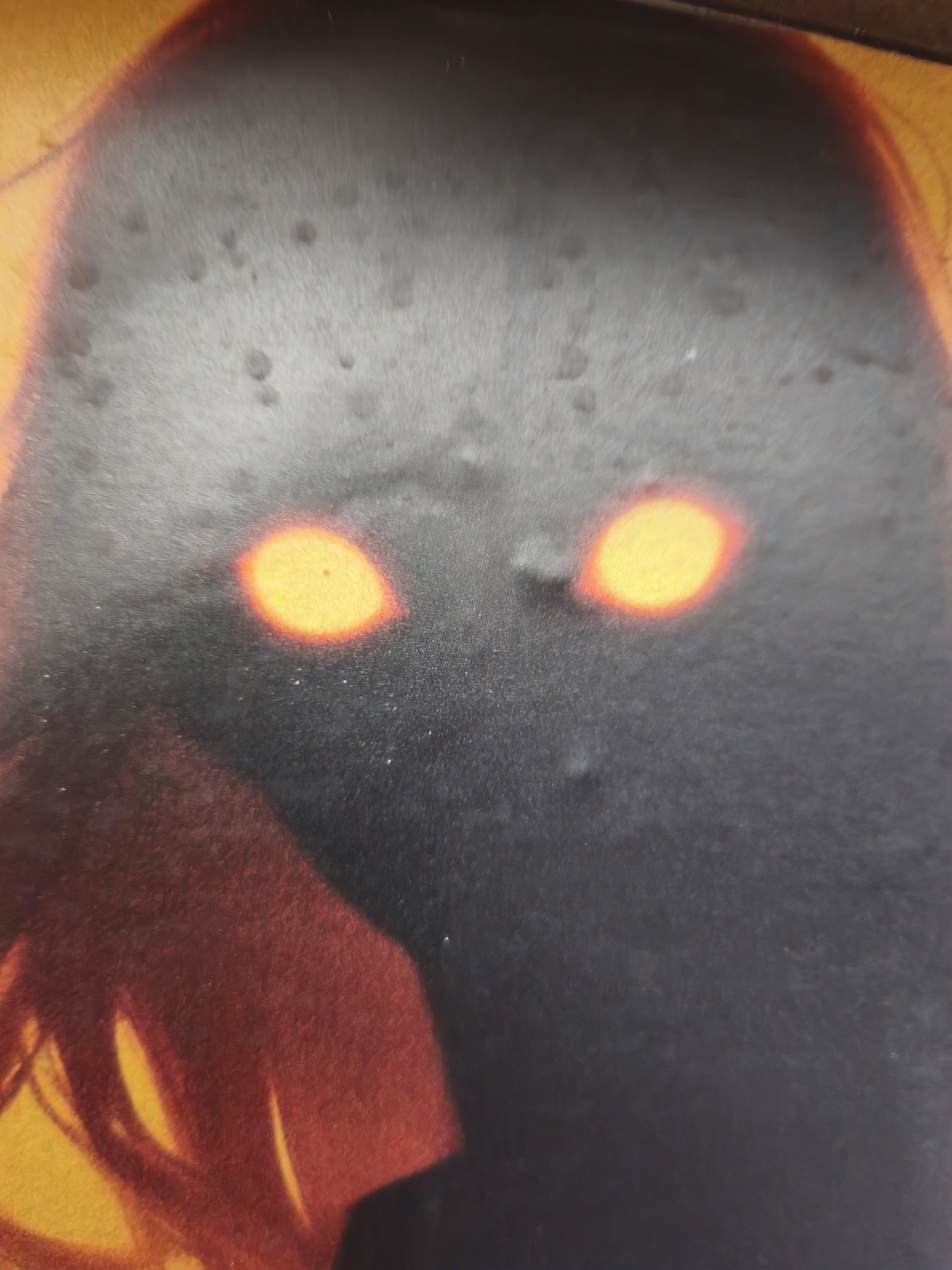 The media file is a close-up, high-angle, slightly blurry photograph of what appears to be a cover or illustration. The dominant subject is a shadowy, humanoid figure with two prominent, glowing orange eyes. The figure's head is large and rounded, and its body is mostly obscured by darkness, with hints of red and yellow light emanating from the lower left side, suggesting a cloak or some other garment. The background is a warm, blurry yellow or orange, with a curved line of red transitioning into a darker shade.
The overall impression is one of mystery and perhaps a touch of menace due to the glowing eyes. There is no discernible activity or event taking place; the image is a static depiction. Notable details include the intense orange glow of the eyes, which stand out against the dark figure, and the slightly textured or speckled appearance of the dark areas, possibly due to the printing or the material of the cover. The lighting appears to be dramatic, with the glowing eyes as the primary light source on the figure. The location context of Kyiv, Ukraine, does not provide any specific visual cues within the image itself. There is no visible text in the media file.