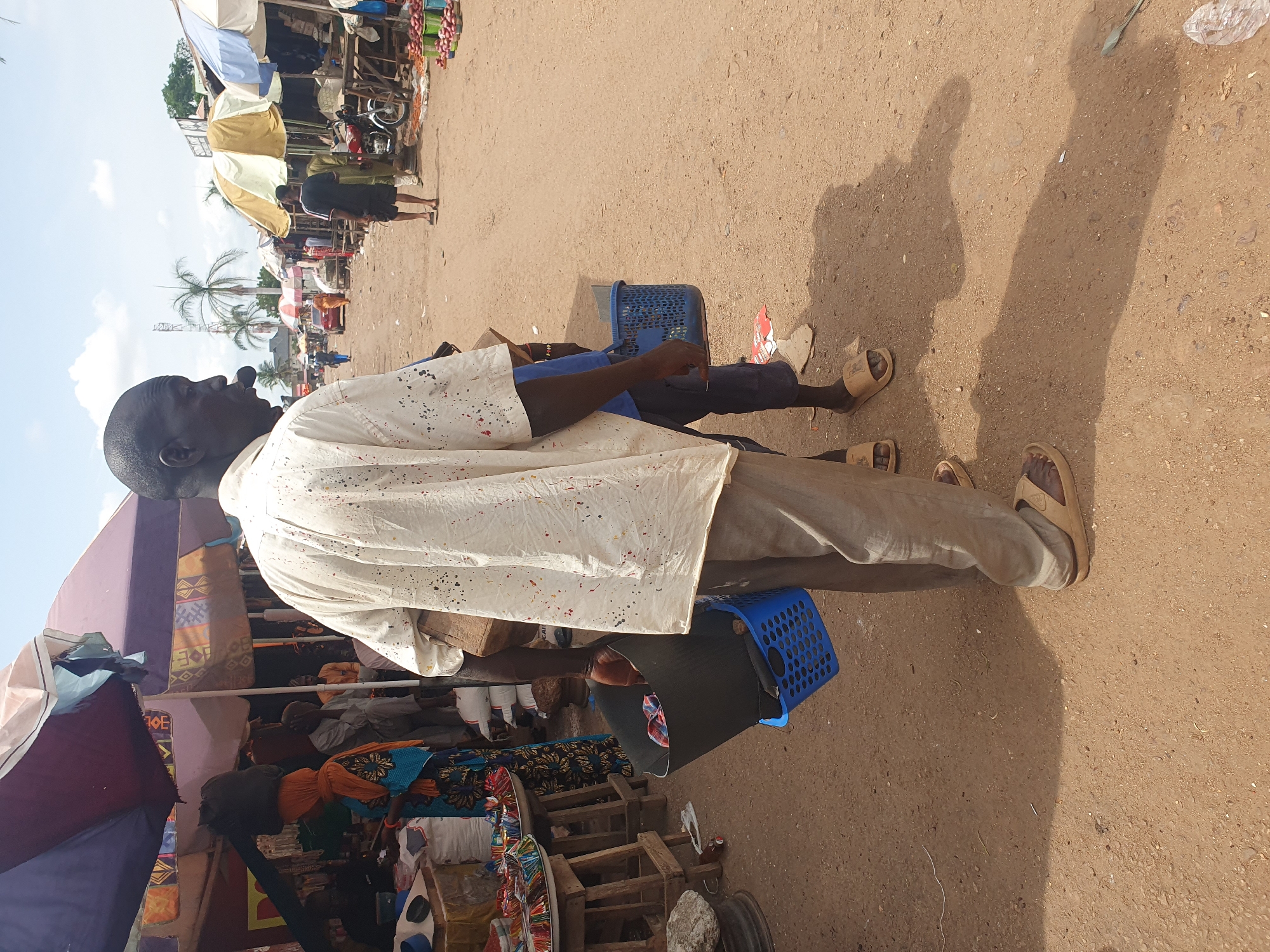 A street view in Lokoja, Nigeria. An African man stands sideways, facing left, wearing a paint-splattered white shirt and tan pants, holding a blue basket. A second blue basket sits on the ground nearby. He wears tan sandals. Other people are visible in the background, along with market stalls shaded by umbrellas and canopies. The ground is dirt.