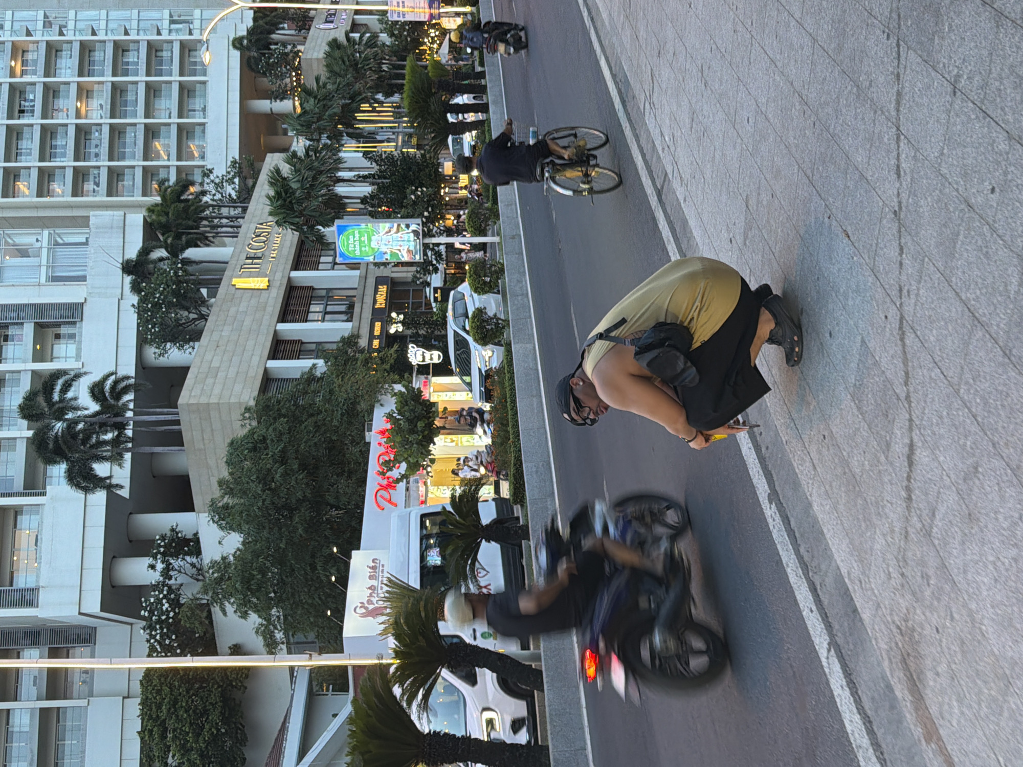 In Nha Trang Ward, Vietnam, a man is squatting on a sidewalk, facing away from the camera. He is wearing a yellow tank top, black pants, and black Crocs. A black bag is slung over his shoulder. Behind him, a busy street is visible with traffic including a motorcycle, a bicycle, and cars. A large modern building with multiple balconies and palm trees lines the street. The overall scene appears to be a snapshot of daily life in a bustling urban environment.