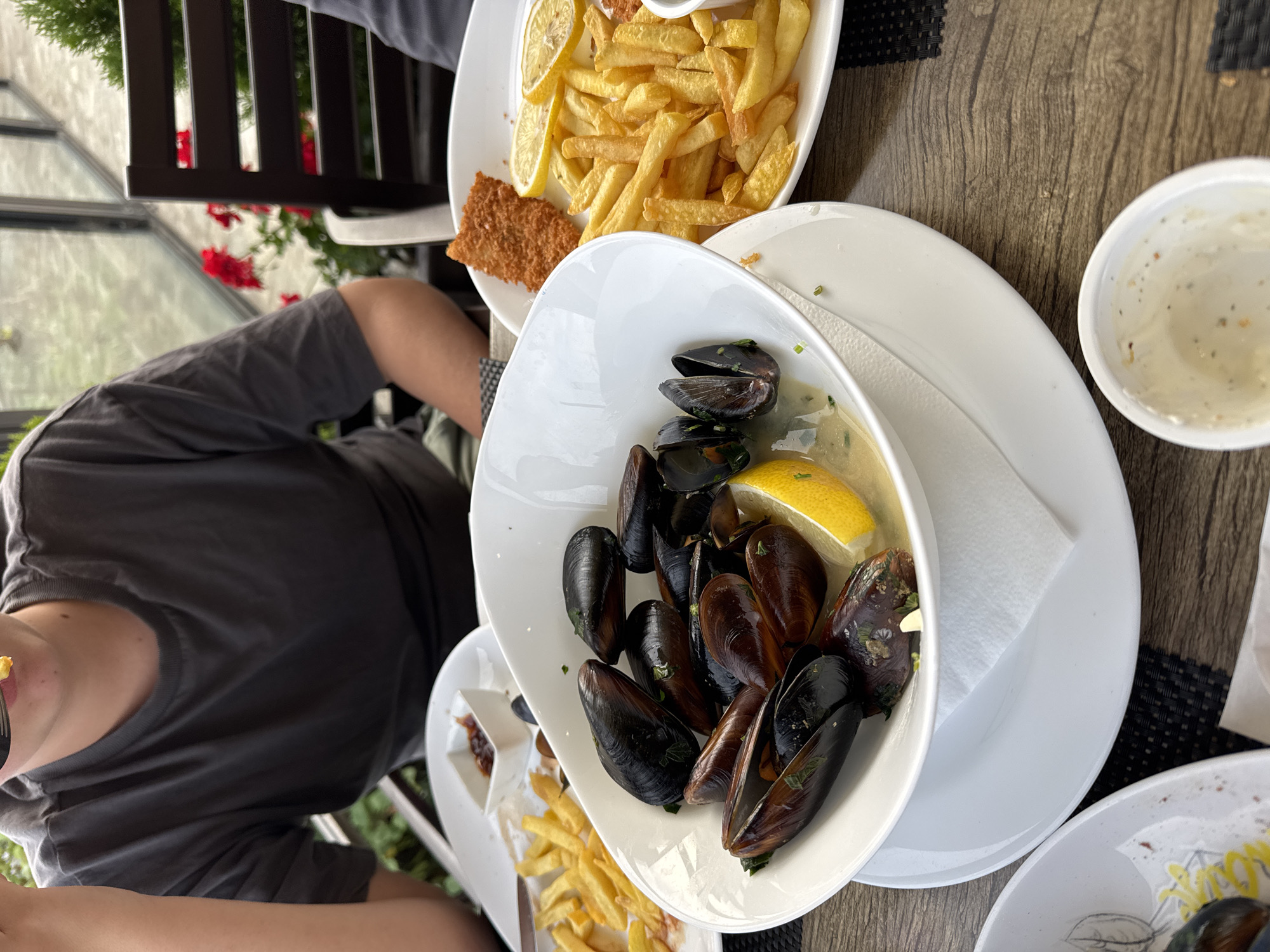A close-up, high-angle shot shows a meal on a table at a restaurant in Sozopol, Bulgaria. In the foreground, a white bowl is filled with mussels, some open and some closed, in a light sauce with a lemon wedge. A white napkin sits beneath the bowl. To the right, a plate holds french fries and a piece of fried fish, garnished with lemon slices. In the background, a person wearing a grey t-shirt is visible, with a fork to their mouth, suggesting they are eating. The table is set with white plates and cutlery. The setting appears to be an outdoor dining area, with blurred greenery and railings visible through a glass partition. The overall atmosphere is casual, with focus on the food.