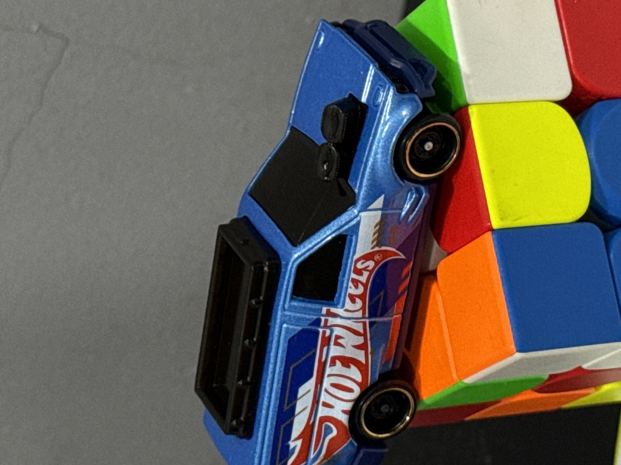 This close-up shot features a vibrant blue toy car, specifically a Hot Wheels model, artfully propped against a multicolored Rubik's Cube. The car is positioned on its side, leaning against the cube, suggesting a playful arrangement of toys.
The toy car is metallic blue with black details on its roof and window frames. Two black wheels with distinctive gold or bronze-colored rims are visible. A prominent "HOT WHEELS" logo in red, white, and blue graphics runs along the side of the car, with the word "HOT" stacked above "WHEELS." The design of the car appears to be a van or station wagon style.
The Rubik's Cube, serving as a prop, displays several colorful faces including white, green, yellow, red, orange, and blue squares. The cube is partially visible, providing a vibrant, geometric backdrop for the toy car. The background is a plain, dark grey, slightly textured surface, keeping the focus entirely on the toys. There are no people, explicit location cues from Da Nang, Vietnam, or discernible time of day or weather conditions present in the image. No emotions are conveyed by the inanimate objects.