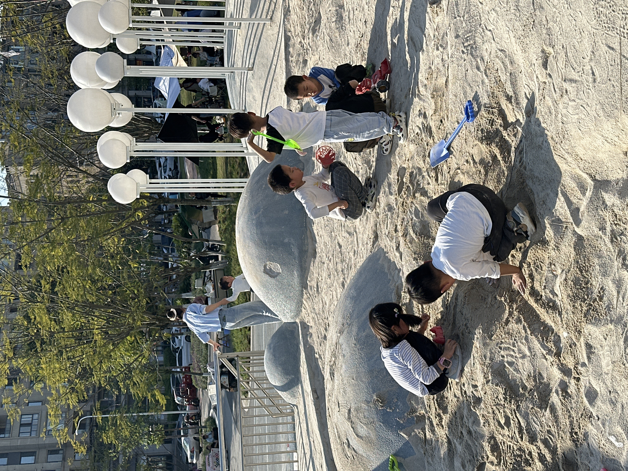 The image depicts a group of children playing in a sand pit at an outdoor park. Five children are visible, ranging in age from approximately 6 to 10 years old. They are interacting with the sand and each other, using toys like shovels and small containers. The sand pit is situated next to a sculptural element made of large, smooth grey rocks.
The backdrop includes trees, a building with multiple stories, and a row of white globe-shaped lamps. A railing separates the sand pit area from a walkway or road where pedestrians are visible. Vehicles are also visible in the background, suggesting an urban setting.
While the image's specific location within the city of Jiangjin, China, is not discernible from the photograph, the presence of modern urban elements confirms the scene's setting is in an urban park.