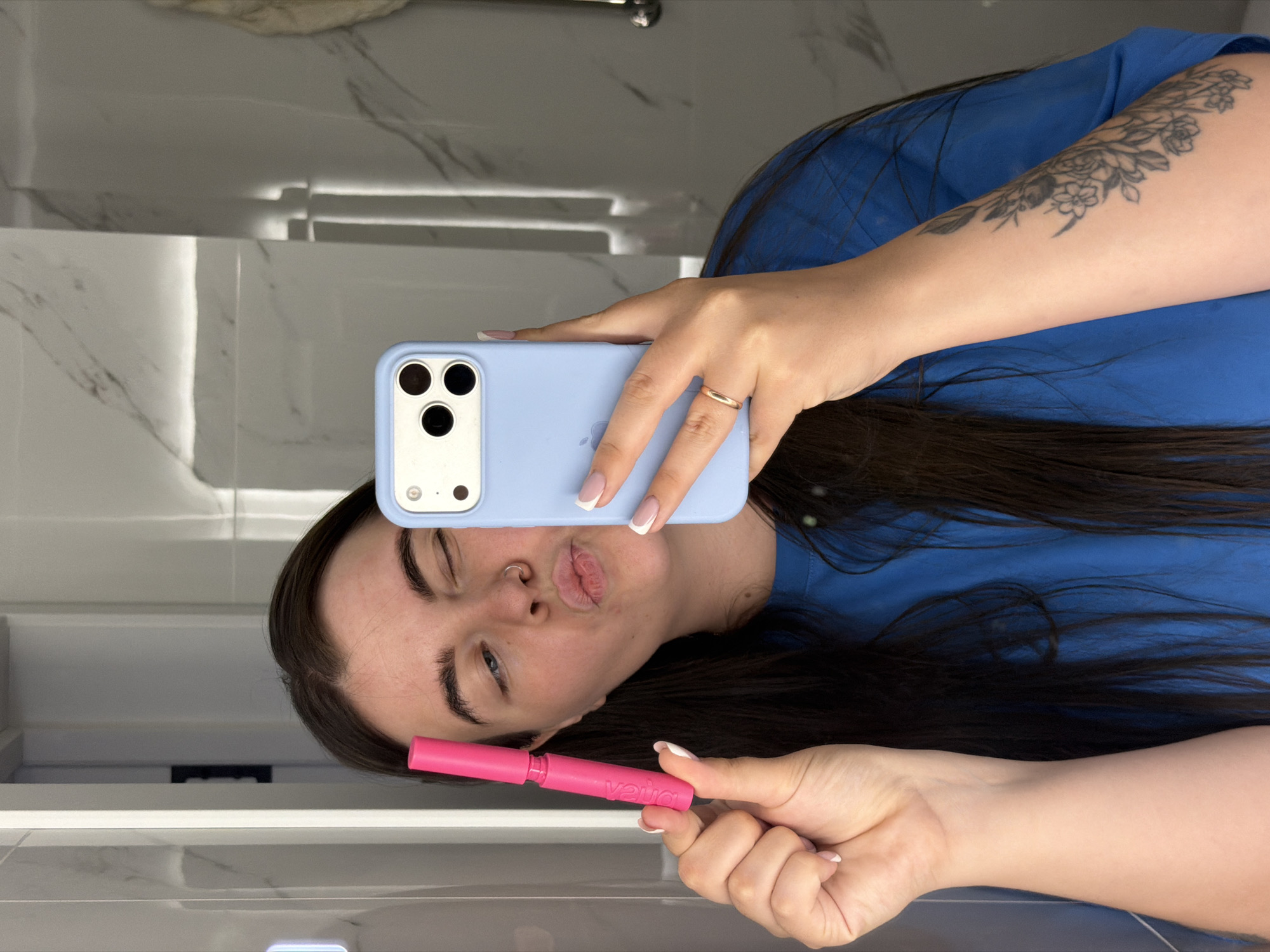A person is holding a blue smartphone in a light blue case with the front camera facing the viewer while standing in front of a mirror in what appears to be a bathroom. The person is wearing a blue t-shirt and has long, dark hair. They are pouting their lips towards the camera. In their other hand, they are holding a pink object, potentially a makeup product or cosmetic item. The background shows a bathroom setting with marble walls and a reflective surface, possibly a vanity or countertop below the mirror. There is no visual confirmation that this image was taken in the city of Chaikovsky, Russia.