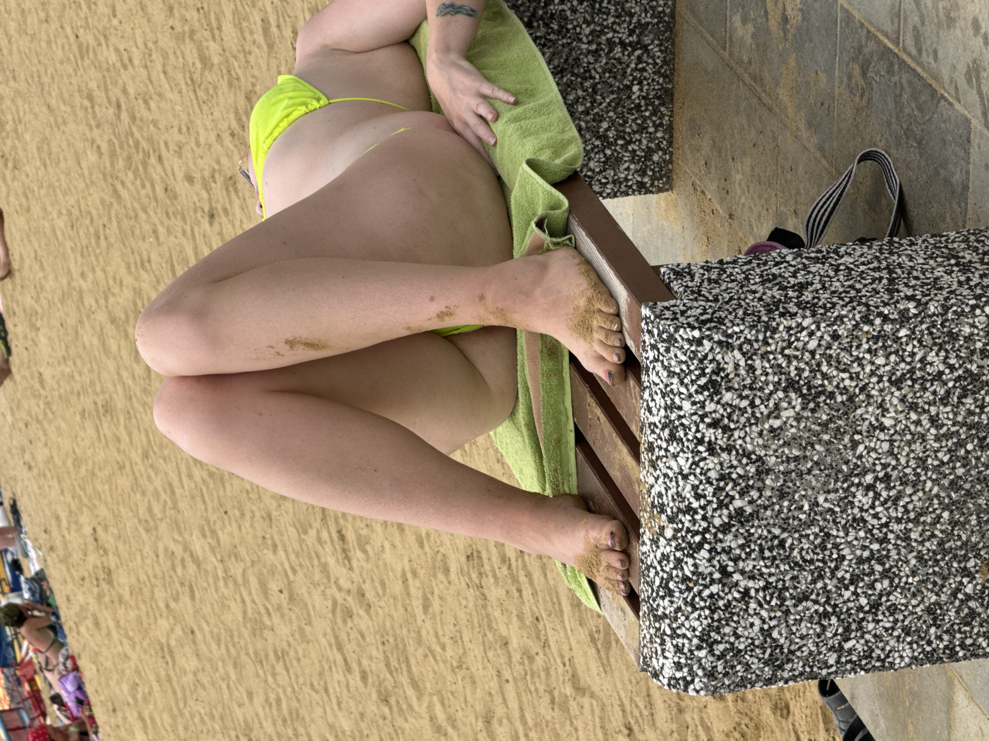 A person lies on a lounger on the sandy beach of Lake Yarovoye in Yarovoye, Russia. The person is wearing a bright yellow bikini. Their legs are propped up, and they are covered in sand. A green towel is laid out on the lounger. The lounger has a speckled stone base. In the background, other people are visible on the beach, some lying on towels or sitting. The water of the lake is not visible in this frame.