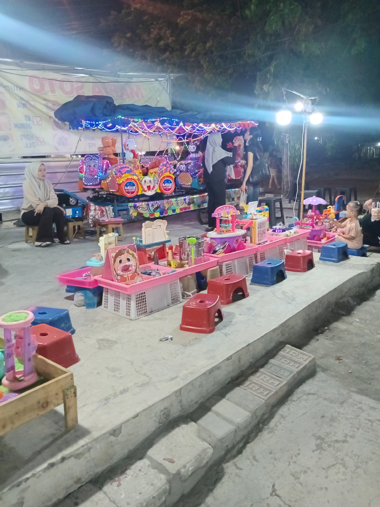 The image depicts an outdoor scene at night, likely a market or a fair, in Sedati Agung, Indonesia. In the foreground, a raised concrete platform is visible, with several colorful plastic stools and low tables displaying children's toys. The toys include miniature kitchens, toy medical kits with cartoonish dental images, and sand play sets. The lighting is artificial, with some areas brightly illuminated by overhead lamps and string lights, while others are in shadow.
In the mid-ground, a small amusement ride with brightly colored carriages decorated with cartoon characters and lights is set up. A woman wearing a hijab is seated on a wooden stool next to the ride, observing the surroundings. Another woman stands nearby, facing away from the camera.
In the background, trees and foliage are visible, suggesting an outdoor setting. A large banner with text, partially obscured by the ride's structure, is stretched across the top of the scene, indicating a commercial establishment or event. The overall impression is of a lively, nighttime gathering, possibly a local festival or a weekend market.