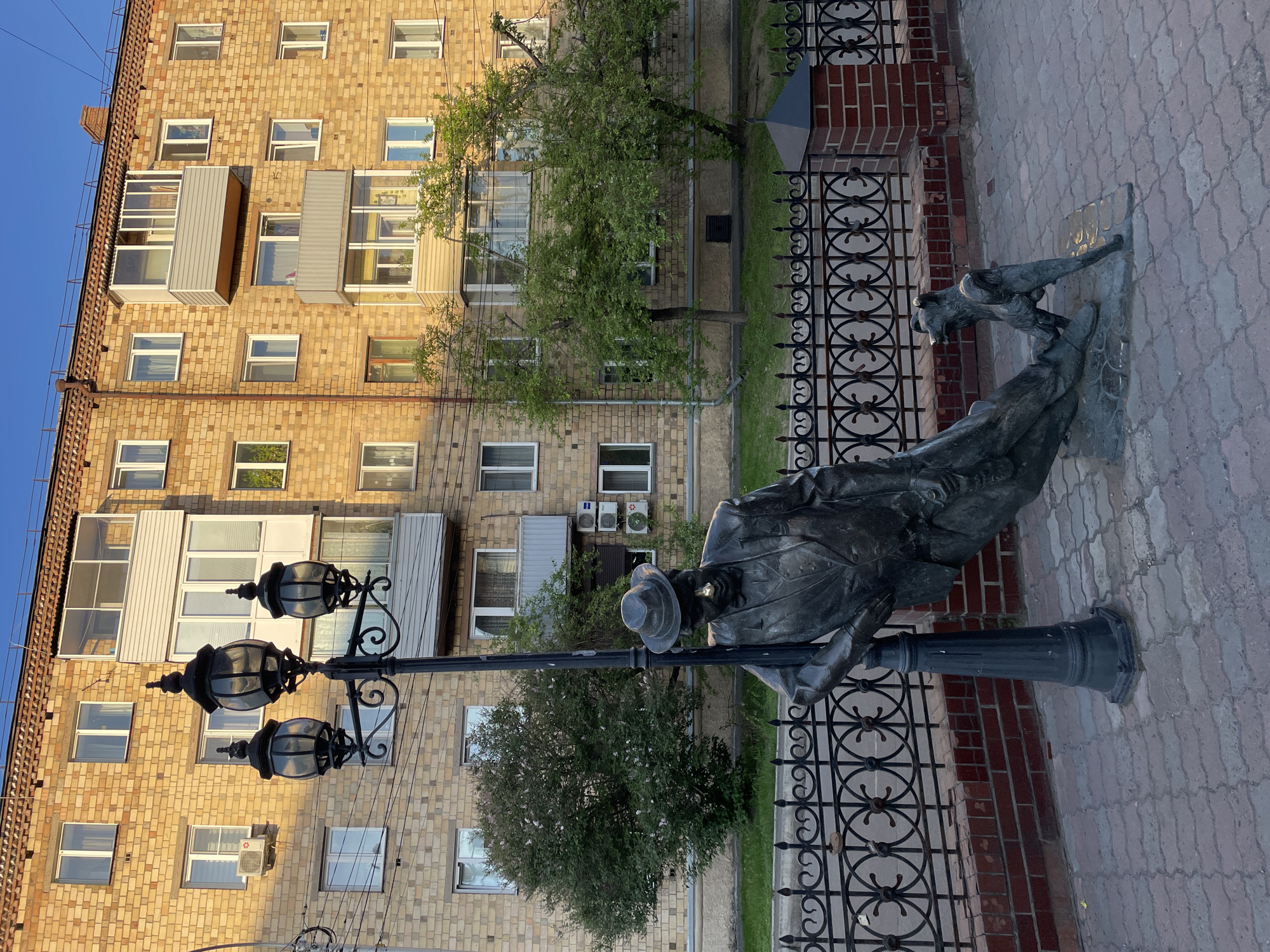 An outdoor urban scene captures a bronze sculpture in the foreground, set against a backdrop of a large brick building and trees. The sculpture, positioned on the right, depicts a man in a hat and coat leaning casually against an ornate lamppost. A dog, standing on its hind legs with paws on the man's leg, gazes up at him, suggesting a moment of companionship or interaction.
A tall, dark, three-lantern lamppost stands prominently, central to the foreground. A decorative black metal fence separates a paved sidewalk from a grassy strip, behind which a low brick wall is visible. The sidewalk is made of grey interlocking pavers.
In the background, a multi-story residential building with a light-colored brick facade features numerous windows, many with white frames, and various balconies, some of which are enclosed. Several air conditioning units are mounted on the building's exterior. Green trees with lush foliage are scattered throughout the mid-ground, providing a natural element to the urban setting.
The lighting indicates a clear, sunny day, likely late afternoon or early morning, with the low sun casting distinct shadows and illuminating the brickwork with a warm glow. The sky is bright blue. No discernible text is visible on any object. The scene reflects an urban environment, consistent with a city like Krasnoyarsk, Russia.