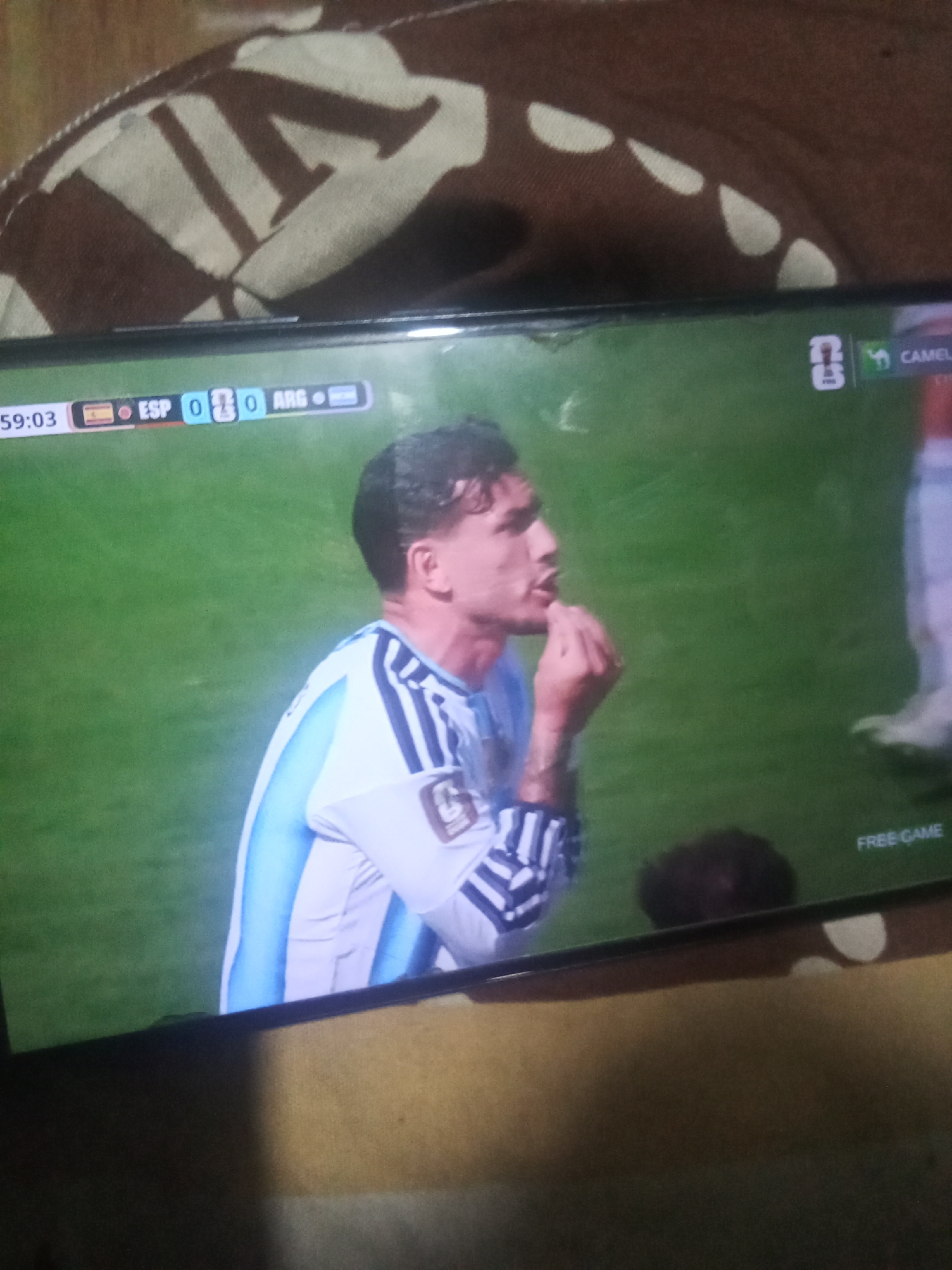 A television screen displays a soccer match. The score shows ESP 0 and ARG 0. A soccer player in an Argentina jersey is kneeling on the field, with his hand on his chin. The background is a green soccer field. The word "FREE GAME" is visible in the bottom right corner of the screen. The image appears to have been taken in a home setting, as a patterned fabric is visible in the background above the television. There is no information to indicate the specific location in Buketglumpang, Indonesia.