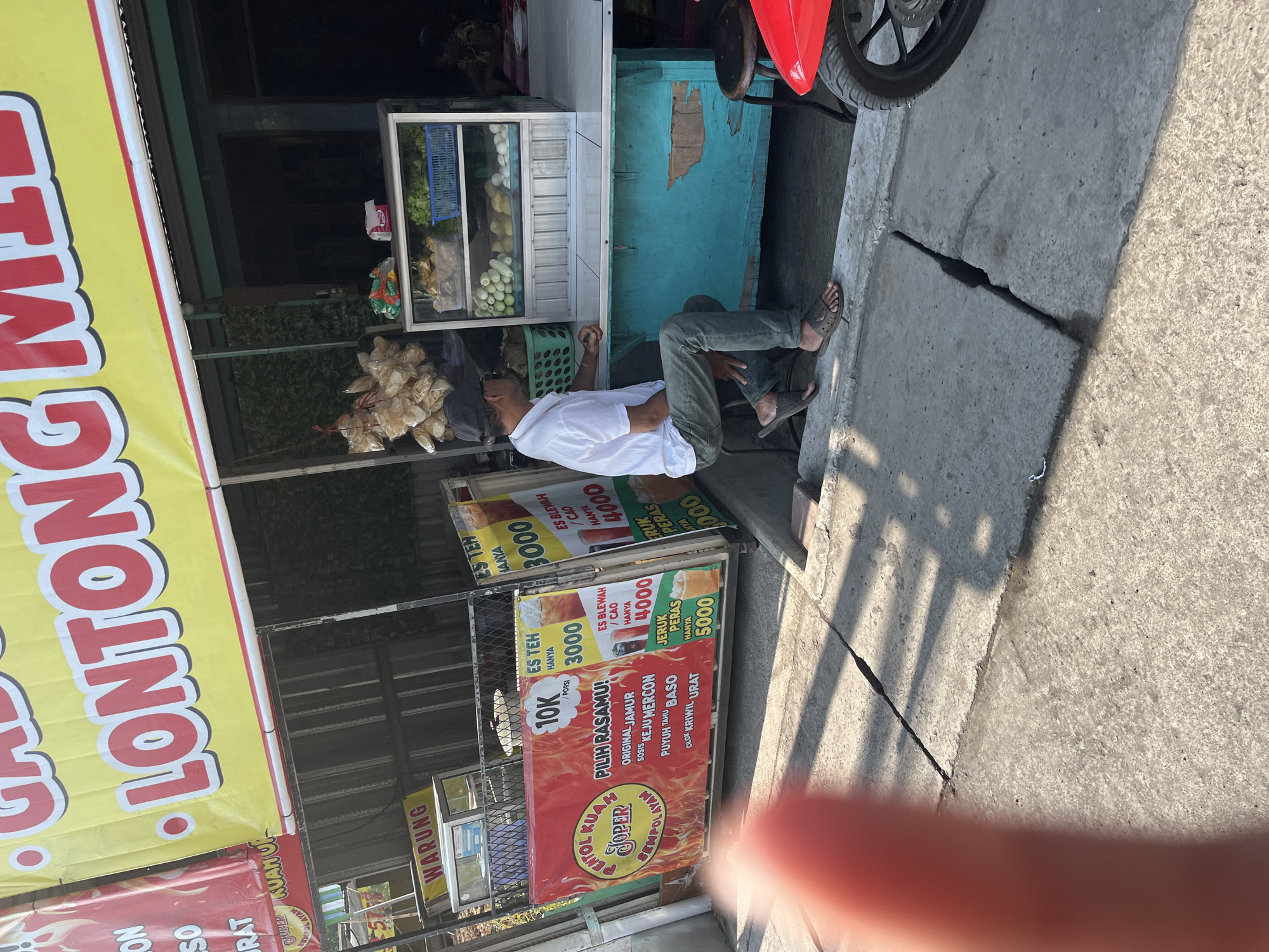 This is a photograph taken outdoors, likely in Tulangan, Indonesia, during the daytime, indicated by the strong shadows cast on the concrete ground. The scene depicts a street food vendor's stall, characterized by a colorful banner that reads "LONTONG." The stall itself appears to be a mobile cart, with a red motorcycle parked nearby.
The primary subject is a man, dressed in a white t-shirt and green pants, seated on a gray concrete slab. He is leaning back and appears to be resting, possibly the vendor or an associate. There are several food items displayed, including what seem to be packaged snacks or crackers in a plastic bag hanging from the stall. A glass display case contains various fresh ingredients, such as vegetables and eggs.
Prominently displayed on the stall are several smaller banners advertising different food and drink items with their prices. These include "ES TEH" (iced tea) for 3000, "ES BLEWAH/CAO" (a type of Indonesian drink) for 4000, and "JERUK PERAS" (freshly squeezed orange juice) for 5000. Another banner with a fiery design advertises "PENTOL KUAH JOPER" with options like "ORIGINAL JAMUR" (mushroom), "SOSIS" (sausage), "KEJU MERCON" (spicy cheese), "PUYUH TAHU BASO" (quail, tofu, meatballs), and "CILOK KRIWIL URAT" (chewy meatballs with veins), all priced at 10,000 per portion. The word "WARUNG" (small shop or stall) is also visible on a sign. The overall impression is a bustling street-side food vending operation in Indonesia.
