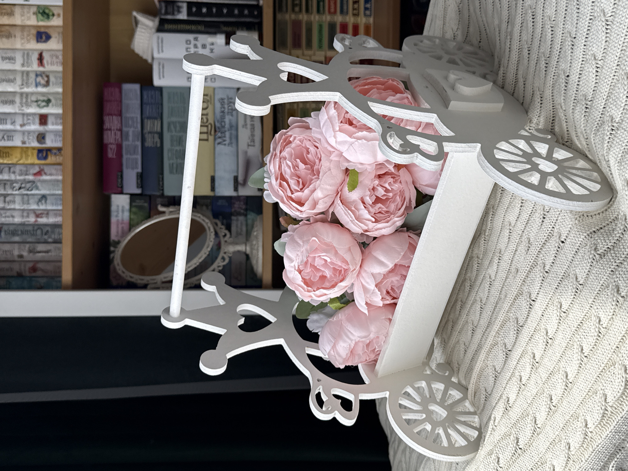 An indoor, medium shot captures a decorative arrangement set against a bookshelf. The image is rotated counter-clockwise. In the foreground, a white, intricately cut wooden or MDF carriage-like object, possibly a decorative sleigh or pram, holds a bouquet of several large, artificial pink flowers resembling peonies. The carriage features ornate cutouts, including heart shapes and crown-like elements, and has decorative wheels on its sides.
To the right, partially visible, is a person wearing a light-colored (beige or cream) knitted garment, possibly a sweater or blanket, with prominent cable-knit patterns, providing a soft, textured background for the carriage.
In the background, on the left, a wooden bookshelf is filled with numerous books arranged vertically. The book spines display various colors, including white, red, blue, green, and yellow, and appear to be in Cyrillic script. Visible titles and authors on some spines include "НОМАНОВЫ" (Romanovs), "ДИКОВ" (Dikov), "ЗВЕРЬ" (Beast), "ИСЧЕЗНУВШИЙ..." (Vanished...), "ЩЕГОЛ" (Goldfinch), and "Мальчик который друг" (The boy who is a friend). Below the main book shelf, a small, ornate, oval-shaped mirror with a white frame is visible on a lower shelf.
The scene depicts a decorative still life within a home environment, likely in Krasnodar, Russia, given the Cyrillic text on the books. The lighting is soft and ambient, suggesting an indoor setting without strong direct light. No specific activity or event is taking place; the focus is on the arrangement of objects.