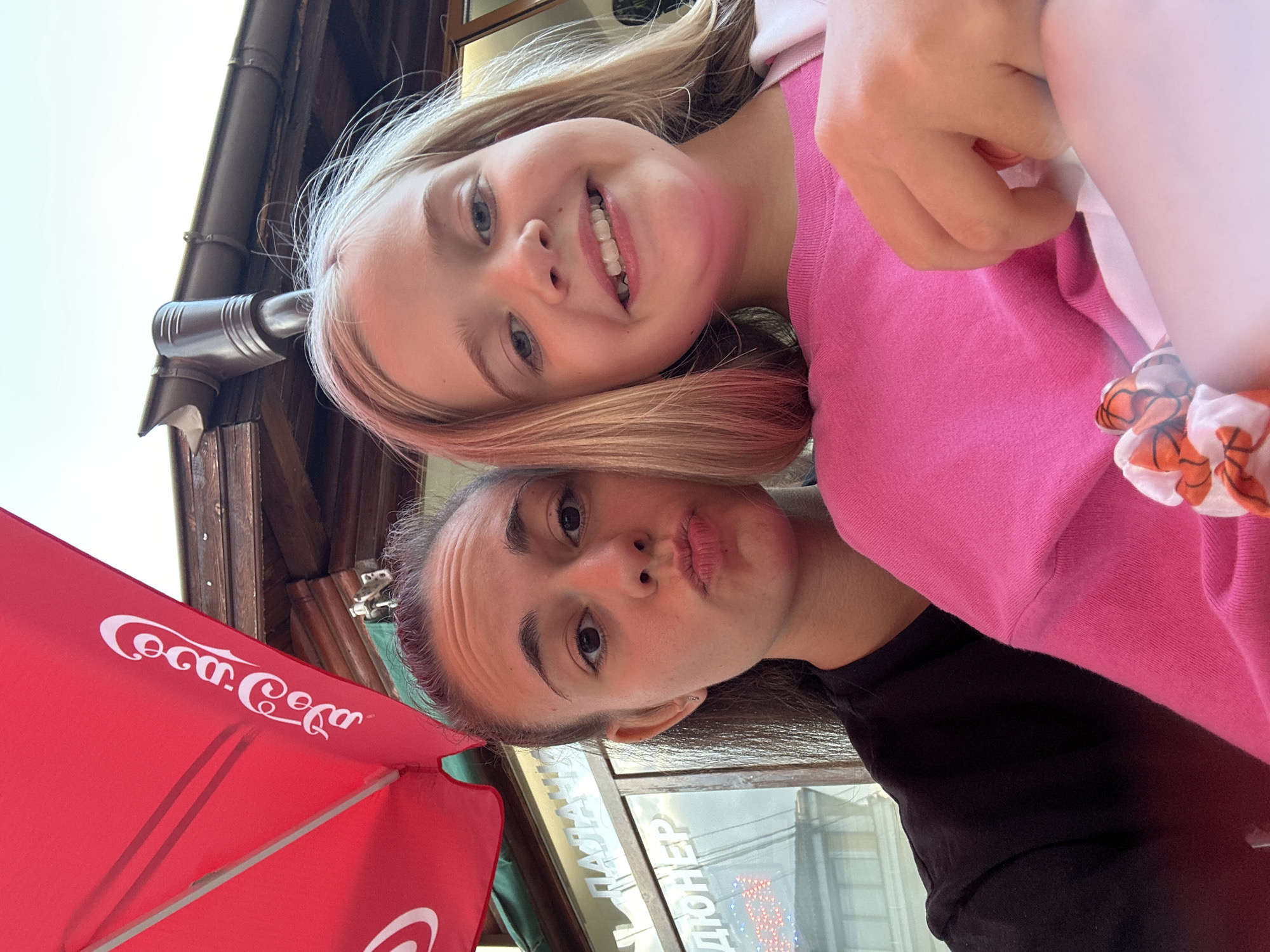 Two young females are captured in a selfie outdoors, under a red Coca-Cola branded umbrella. The female on the left, with dark hair pulled back, is making a kissing face. The female on the right, with blonde hair, is smiling. Behind them, a wooden structure with a gutter is visible, along with signage that appears to be in Cyrillic script, suggesting a location in Samokov, Bulgaria. The scene is casual, likely taken during a daytime outing.