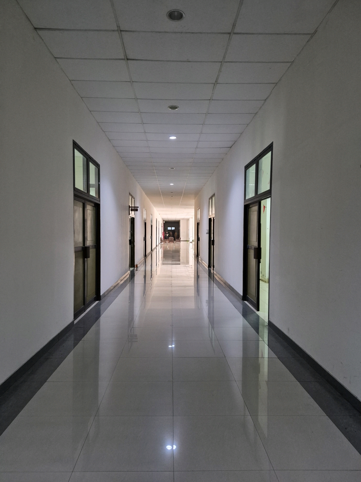 This is a wide-angle, eye-level photograph of a brightly lit, modern hallway with a polished tile floor that reflects the ceiling lights. The hallway extends into the distance, with doors and windows lining both the left and right walls. The walls are painted white, and the ceiling is a grid of acoustic tiles with recessed lighting. The floor is tiled in a light color with dark grout lines, and its reflective surface creates strong reflections of the ceiling and the doors. The scene suggests an institutional or commercial building. The specific location in Purworejo, Indonesia, is not discernible from the image alone, as the architectural style is generic and could be found in many locations.