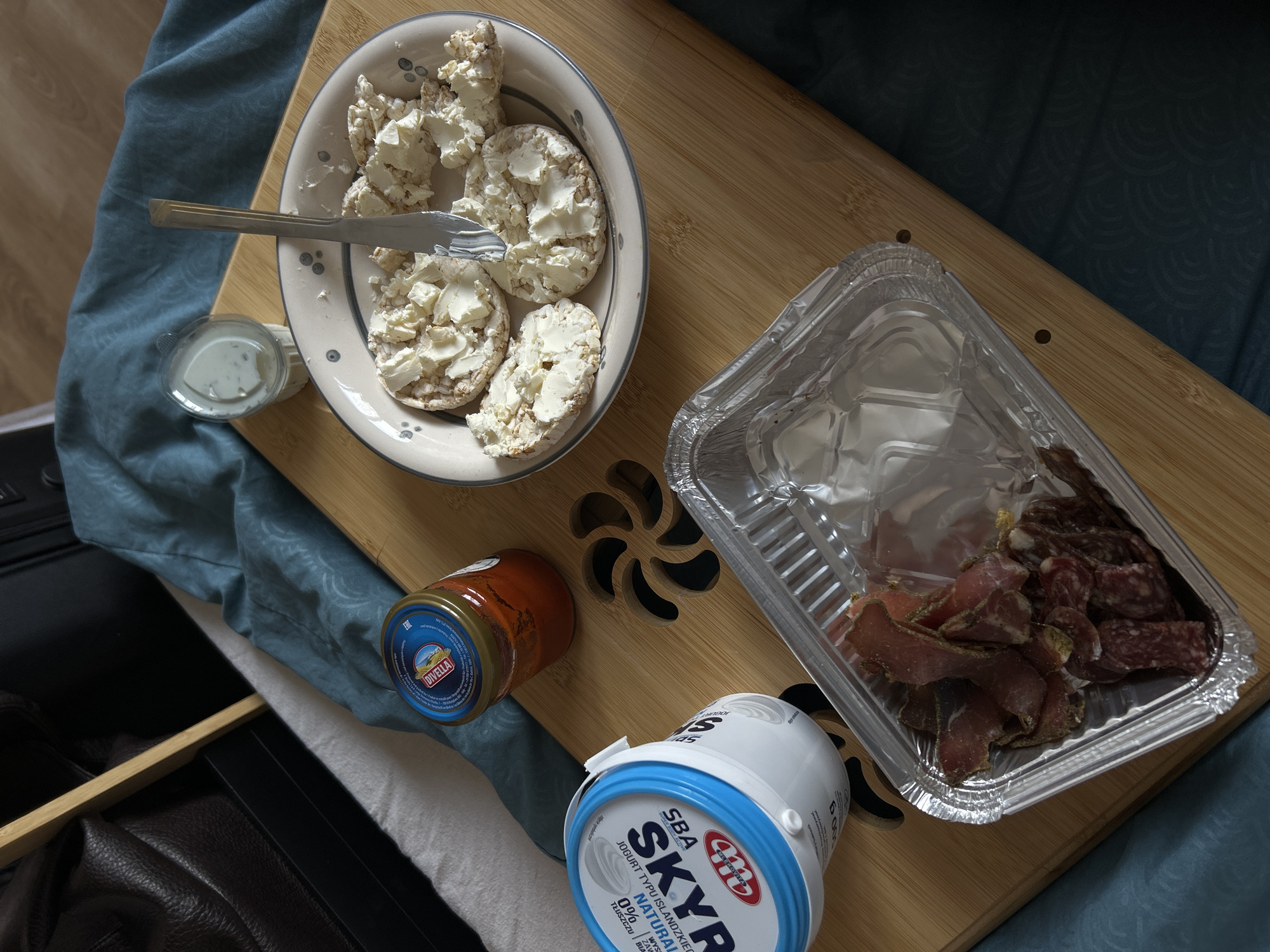 The image shows a meal arrangement on a wooden tray set on a bed covered with a patterned blue fabric. A white bowl contains several rice cakes topped with a creamy white substance, likely cottage cheese. A silver knife rests across the bowl. Next to the bowl is a small plastic container filled with a white sauce.
To the right of the bowl, there is a rectangular foil container holding slices of dried meats, including what appears to be prosciutto and salami. Below the foil container is a jar of "Divella" sauce and a blue and white container of "Skyr" yogurt.
The lower left corner of the image shows a portion of a dark leather or faux-leather object, possibly furniture or a bag.
There is no information available in the image regarding the location in the city of Sofia, Bulgaria, or any people present.