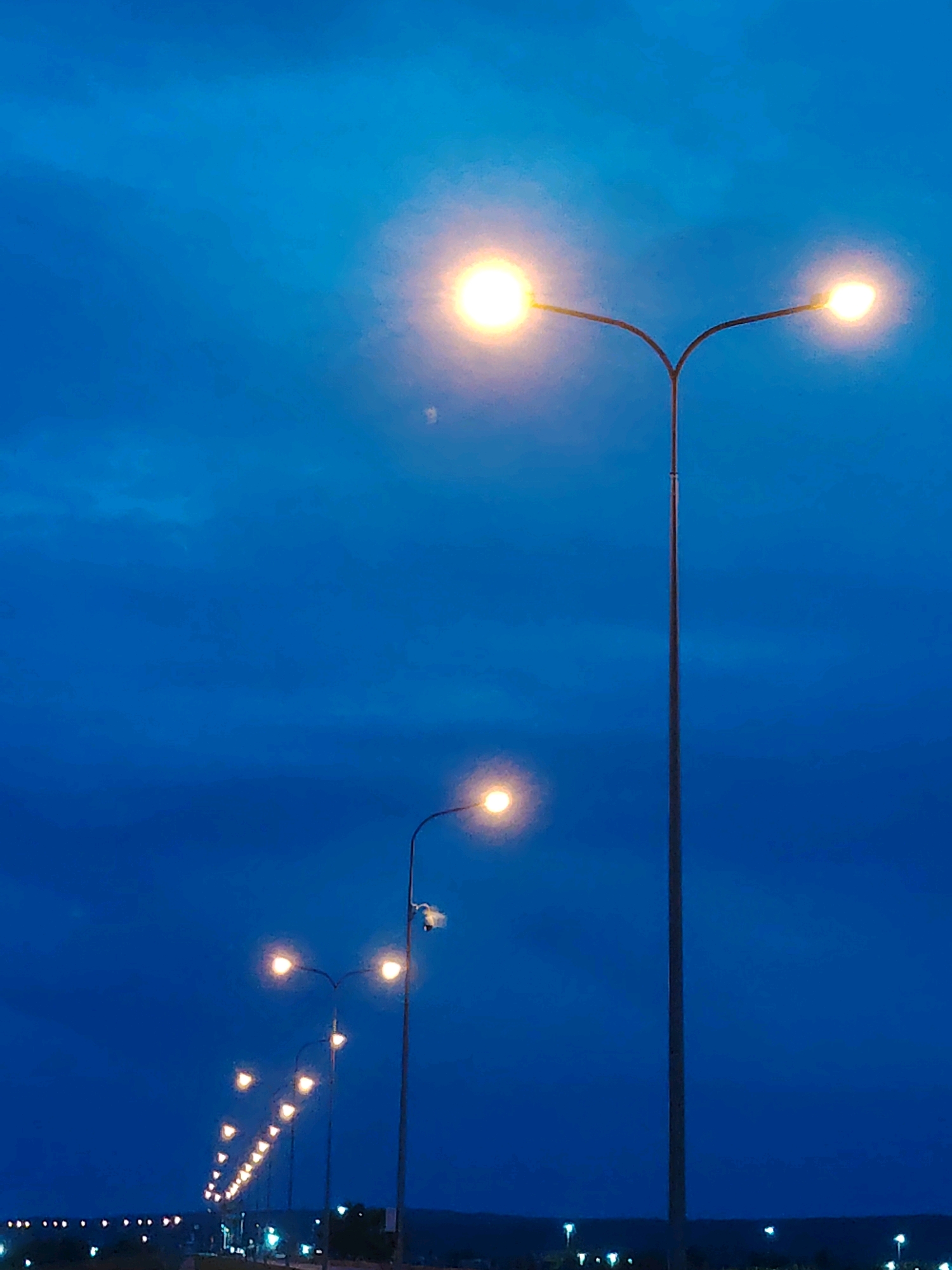 The image depicts a street scene at dusk, likely in the city of Chaikovsky, Russia. A series of streetlights illuminate the scene, with a prominent dual-headed streetlight in the foreground on the right. The sky is a deep, dark blue, indicative of twilight, with subtle cloud formations visible. In the distance, more streetlights form a receding line along what appears to be a road or pathway, and further still, faint lights suggest buildings or structures on the horizon. The overall impression is one of a quiet, transitional moment between day and night.