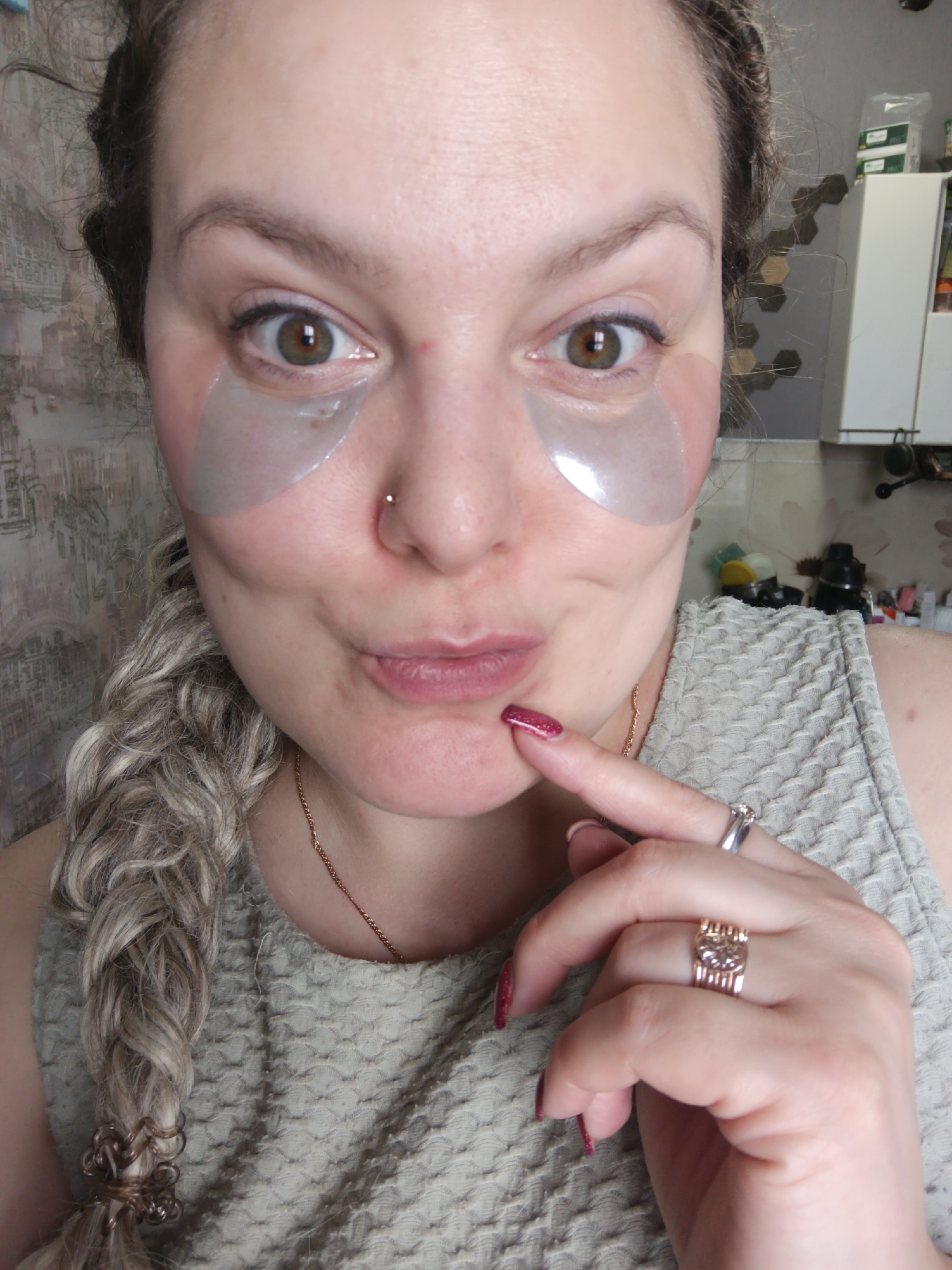 A woman is depicted wearing eye patches, with a nostril piercing visible. Her hair is styled in a braid, secured with decorative accessories. She is wearing a textured sleeveless top. She has rings on her fingers. Her nail polish is red. The location appears to be an interior space with decorative wallpaper. Based on the location provided (Марковский, Russia), the scene is likely within a residential or personal space in that city.