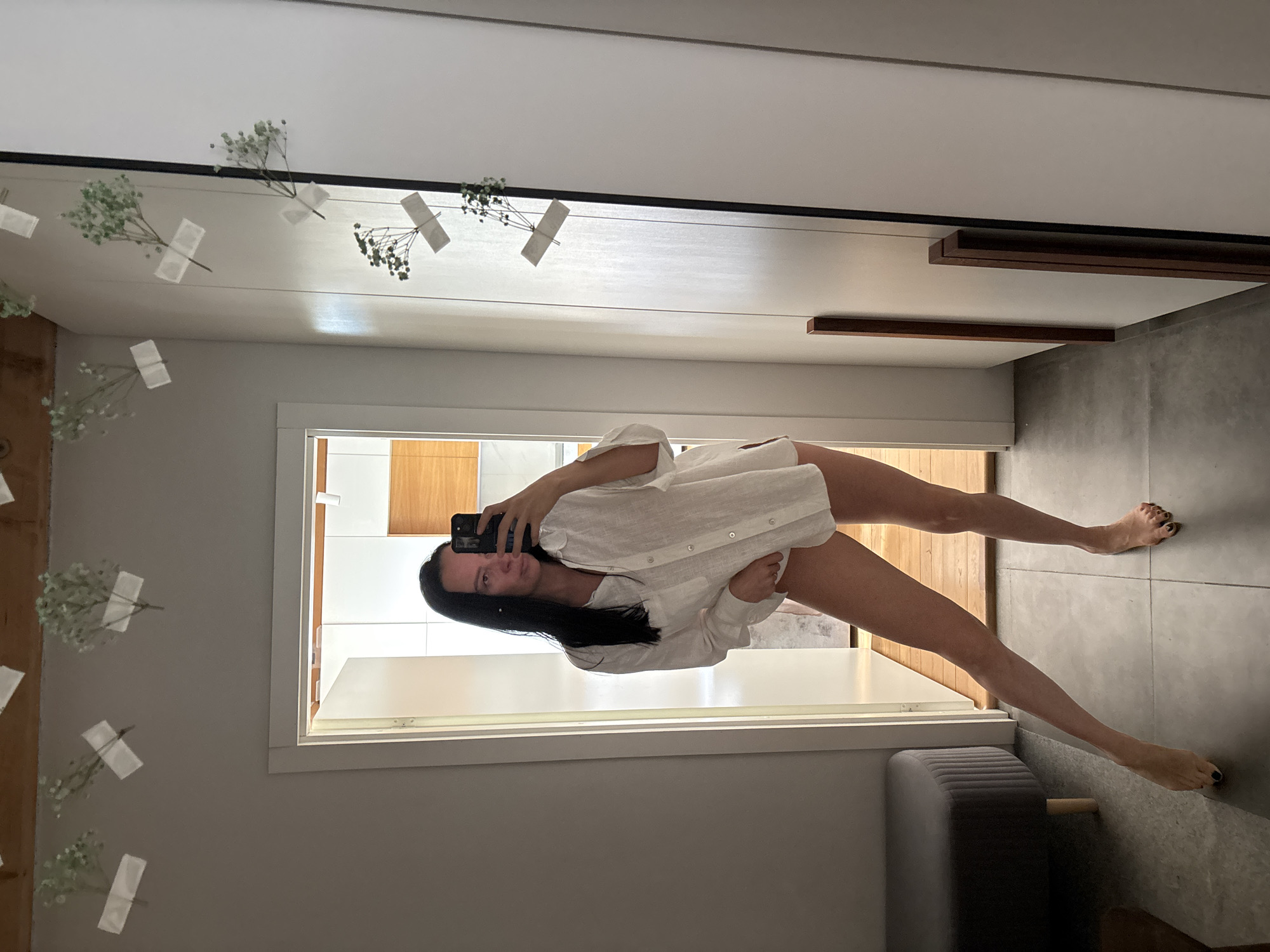 A woman is reflected in a mirror taking a selfie. She has long dark hair and is wearing a white button-down shirt. The background includes a hallway with wood flooring and gray tile. Flowers are taped to the top of the mirror frame. There is also a gray bench visible. The photograph appears to be taken in an interior space and based on the available information, it cannot be determined if the location is in the city of Chaikovsky, Russia.