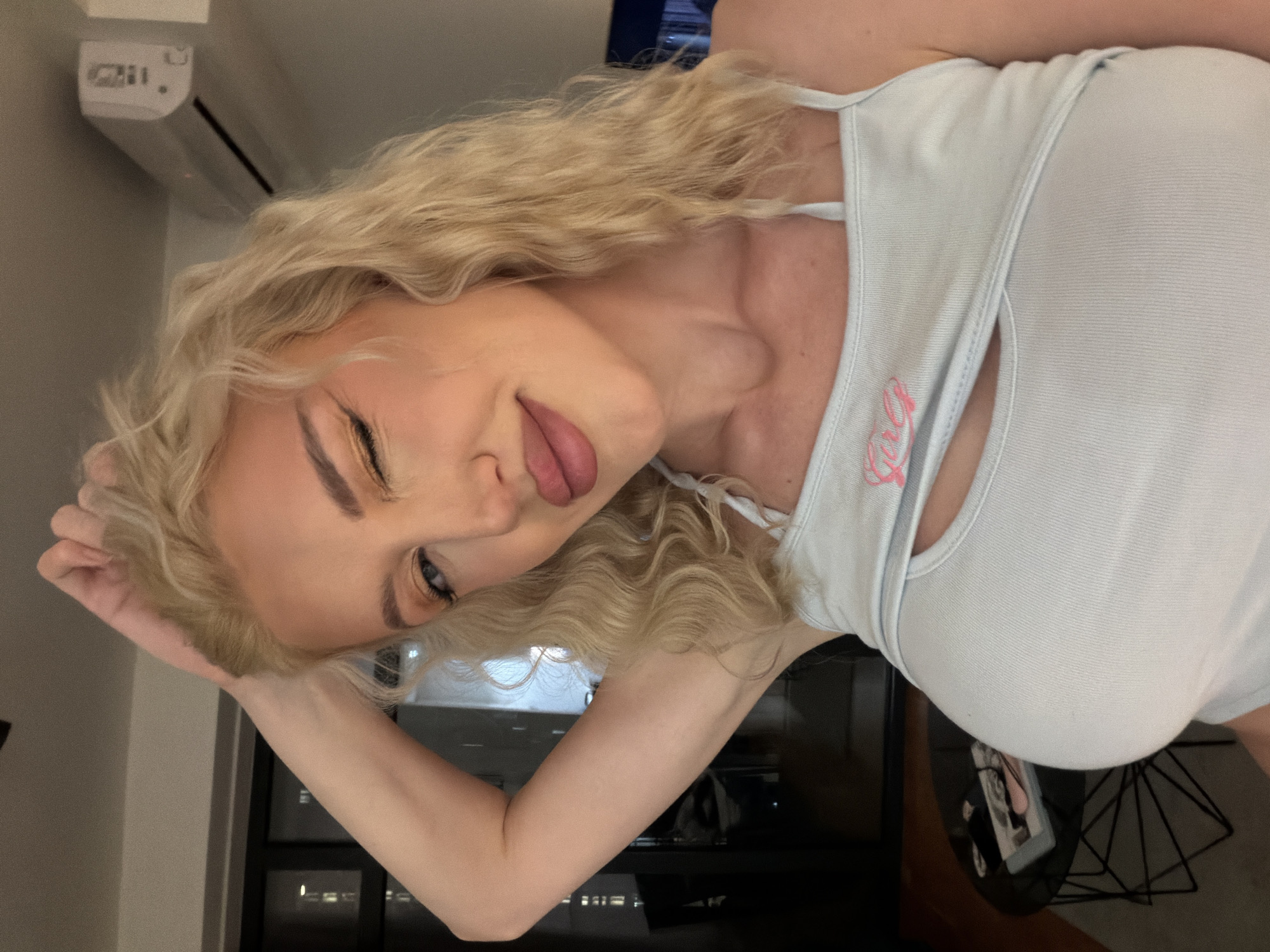 A blonde-haired woman with blue eyes wearing a light blue tank top with pink writing is sticking out her tongue and winking. She appears to be indoors at night, with a window showing city lights in the background. The presence of an air conditioning unit on the wall indicates the image was taken indoors. Although the city is not clearly visible, it may be in Belgrade, Serbia.