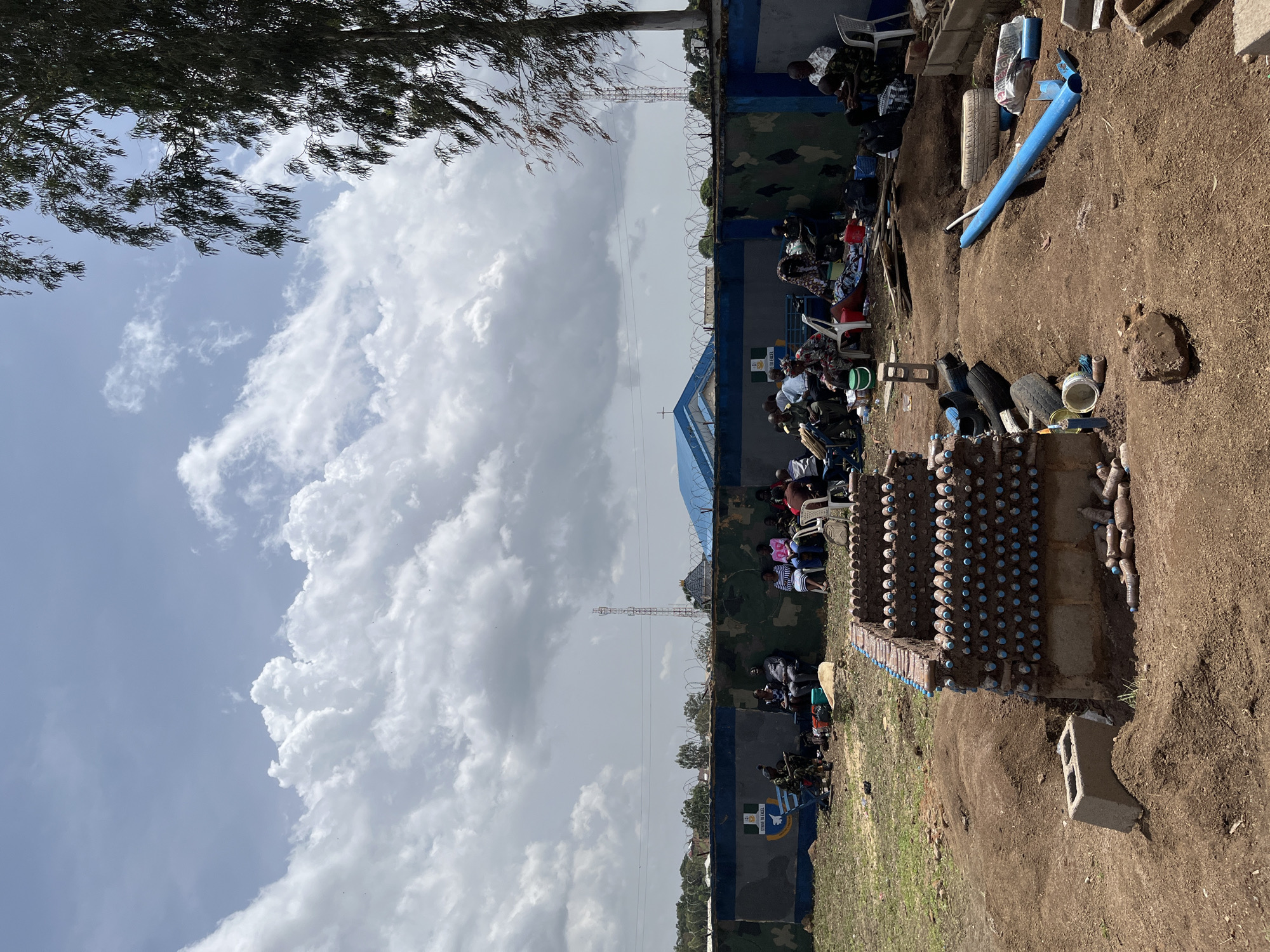 In Bar Jirgi Summa, Nigeria, a group of individuals are gathered outdoors, some seated on chairs and benches, while others are on the ground. They are positioned in front of a wall adorned with a camouflage pattern and an emblem. The wall appears to be part of a fenced area, with barbed wire visible along the top. Behind the wall, a blue-roofed building and a communication tower are discernible. In the foreground, a partially constructed structure made of bottles and mud is present, alongside piles of earth and various objects including tires, pipes, and building materials. The sky above is filled with dramatic clouds.