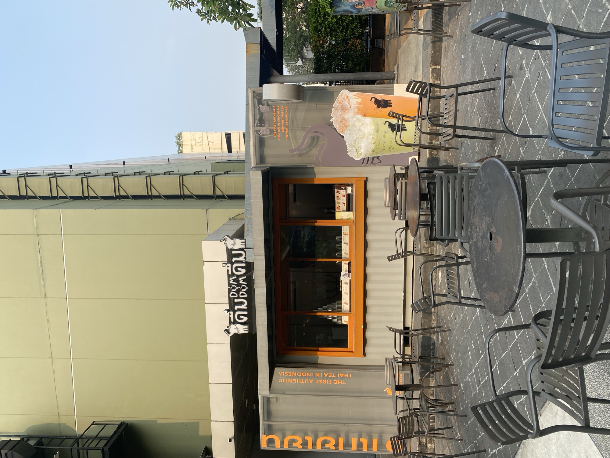 This is an outdoor shot of a coffee shop with a few tables and chairs. The shop is a modern-looking building with a large orange-framed window and a prominent sign. The sign features the shop's name, "ARIRU" in large orange lettering, and a smaller sign with "THE FIRST AUTHENTIC THAI TEA IN INDONESIA". Inside the window, there are rows of stacked cups. The tables and chairs are made of dark metal, and the ground is paved with dark cobblestones. The lighting suggests it is daytime, likely morning or late afternoon, as there are shadows and the sky is bright. The scene is calm and appears to be a quiet moment outside the shop. The overall impression is one of a clean, contemporary establishment ready for customers.
