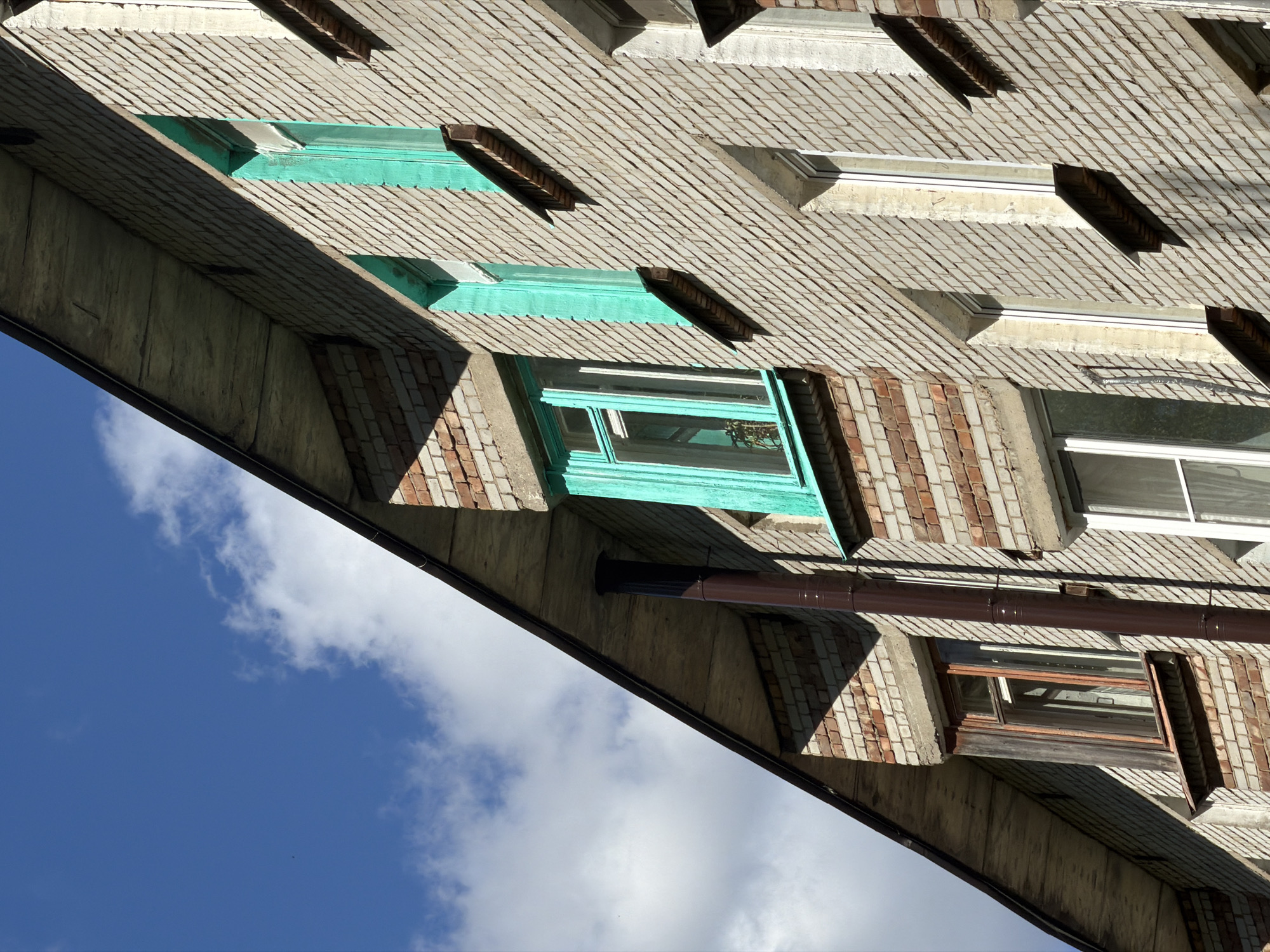 The image shows the exterior of a brick building in Virki-1, Russia, viewed from a low angle pointing upward. The building has multiple windows with varying frame colors including light green and white. The sky is visible in the lower left of the image, featuring blue sky with white clouds. A brown drainpipe runs along the building's facade.