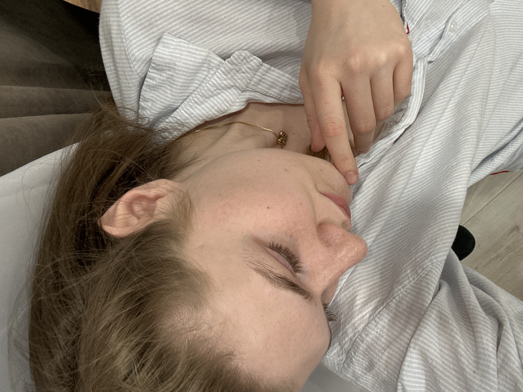 A person with light skin and brown hair lies supine on a white surface, possibly a bed or examination table. They are wearing a light blue and white striped shirt. The person's right hand is resting near their face. A gold necklace with a charm is visible around their neck. The background includes a light brown surface, possibly a wall or furniture, and a section of light-colored flooring. The location in the city of Kazan, Russia, is not discernible from the provided image.