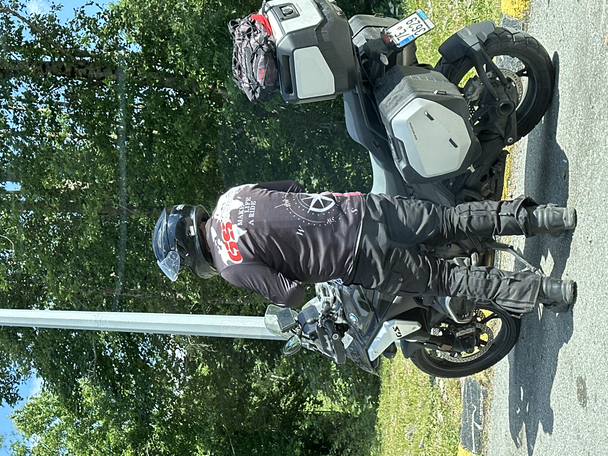 A person wearing a black motorcycle helmet and a black long-sleeved shirt with red and white graphics stands next to a black BMW GS motorcycle. The shirt has the words "MAKE LIFE A RIDE" printed on it, along with a compass graphic. The person is wearing black motorcycle pants and black boots. The motorcycle has two silver panniers and a black top case with a red and black bag strapped to it. A license plate, "7E 3629," is visible on the rear of the motorcycle. The scene appears to be on the side of a road, with trees and greenery in the background. The lighting suggests it is daytime, with sunlight casting shadows. The image was captured in Crkvine, Montenegro.
