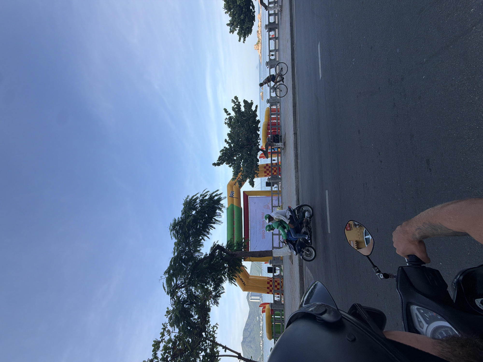 A view from the perspective of someone on a scooter in Nha Trang, Vietnam, looking towards the coastline. In the foreground, the handlebar and a portion of the scooter's dashboard are visible, along with the rider's arm. The road is paved, and a white line marks a lane.
Further ahead, a yellow and green inflatable archway stands on the promenade. Behind the archway, a large white banner with text is displayed. Two people are riding a motorcycle towards the left, both wearing green helmets. To the right of the archway, a person is riding a bicycle along the promenade.
The promenade overlooks the sea, with boats visible in the distance. Mountains form the backdrop, and a city skyline can be seen on the left side. Trees line the promenade, and the sky is a clear blue with some light clouds. The overall scene suggests a public event or gathering is taking place.